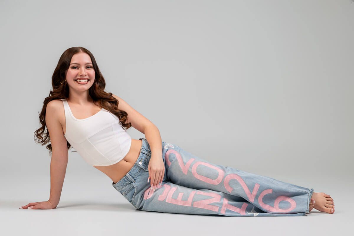 Young woman smiling, wearing white crop top and graphic jeans, posing on floor against gray background.