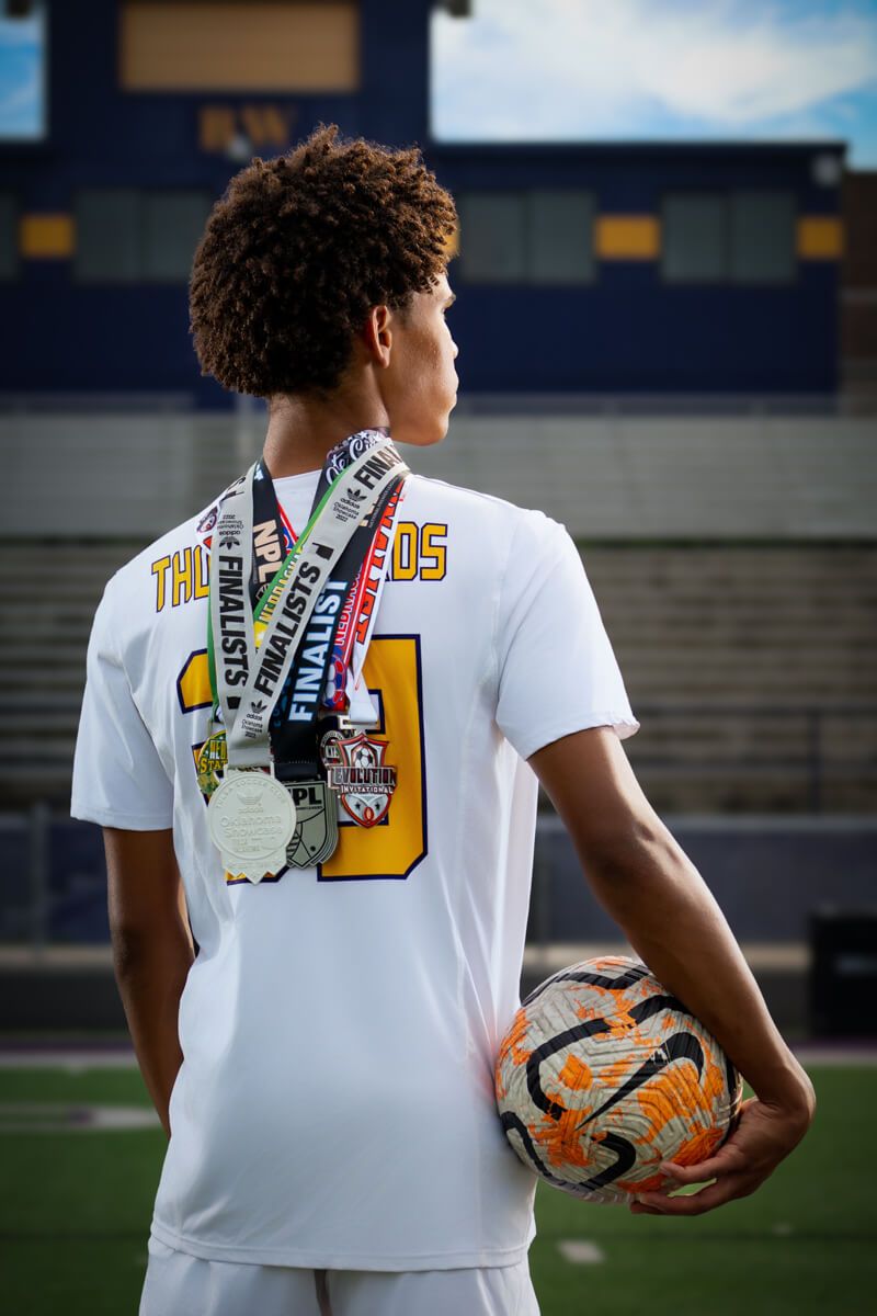 Young soccer player in white jersey holding a Nike ball, wearing finalist medals, standing on a stadium field.