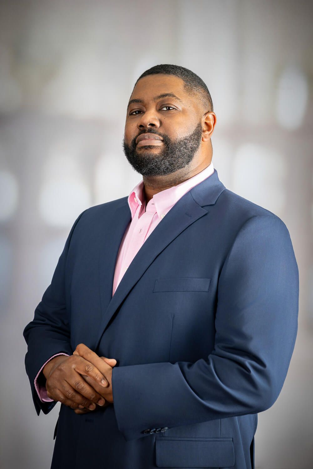 Professional portrait of a person in a navy suit and pink dress shirt against a blurred background.