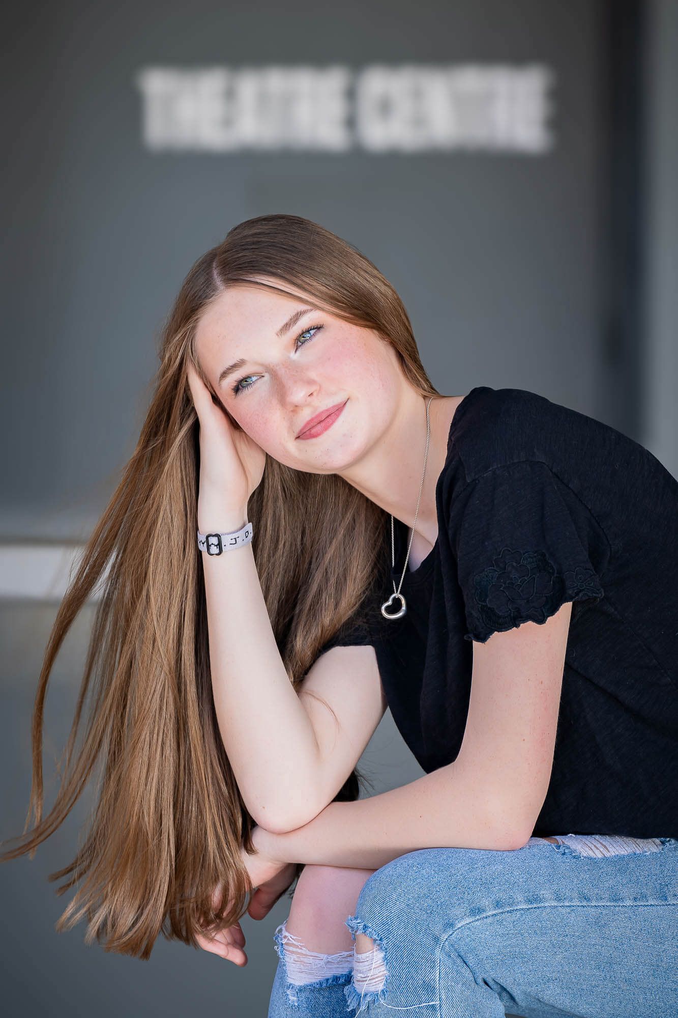 Young woman in black top and jeans sits casually with long brown hair flowing while smiling at camera.