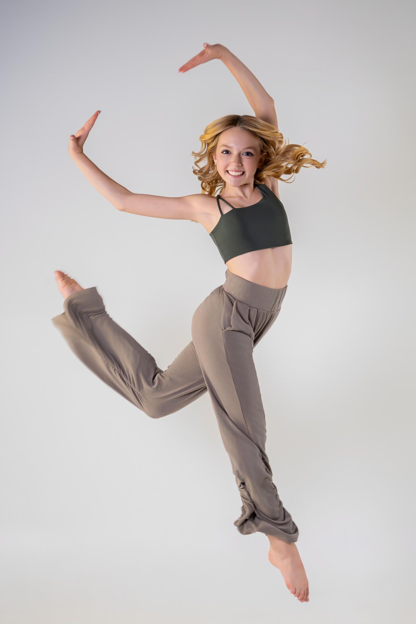 Dancer in black crop top and taupe pants performs dynamic leaping sequence against white studio background.