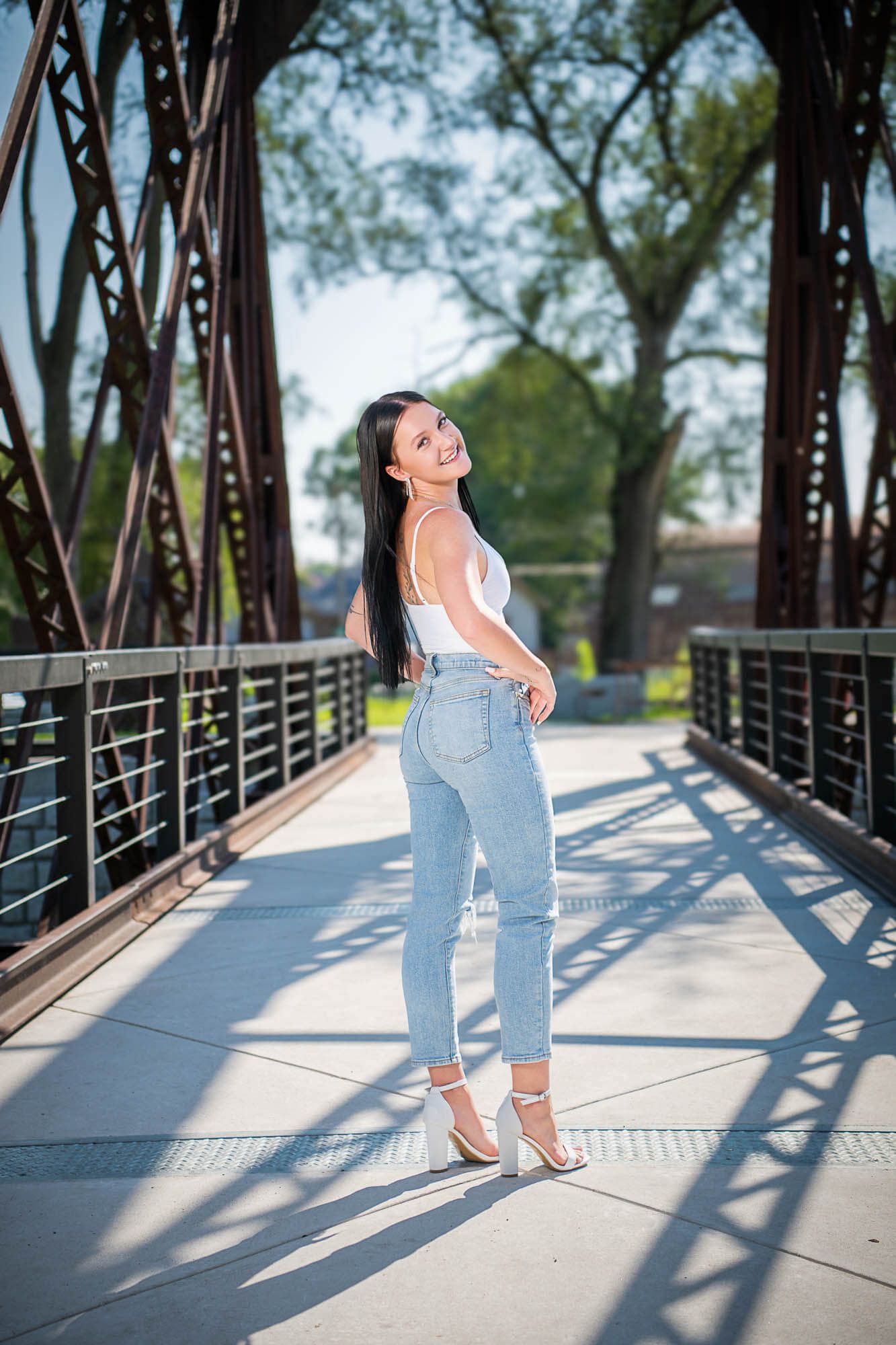 A person poses on a metal bridge walkway wearing light blue jeans, white top and heels on a sunny day.