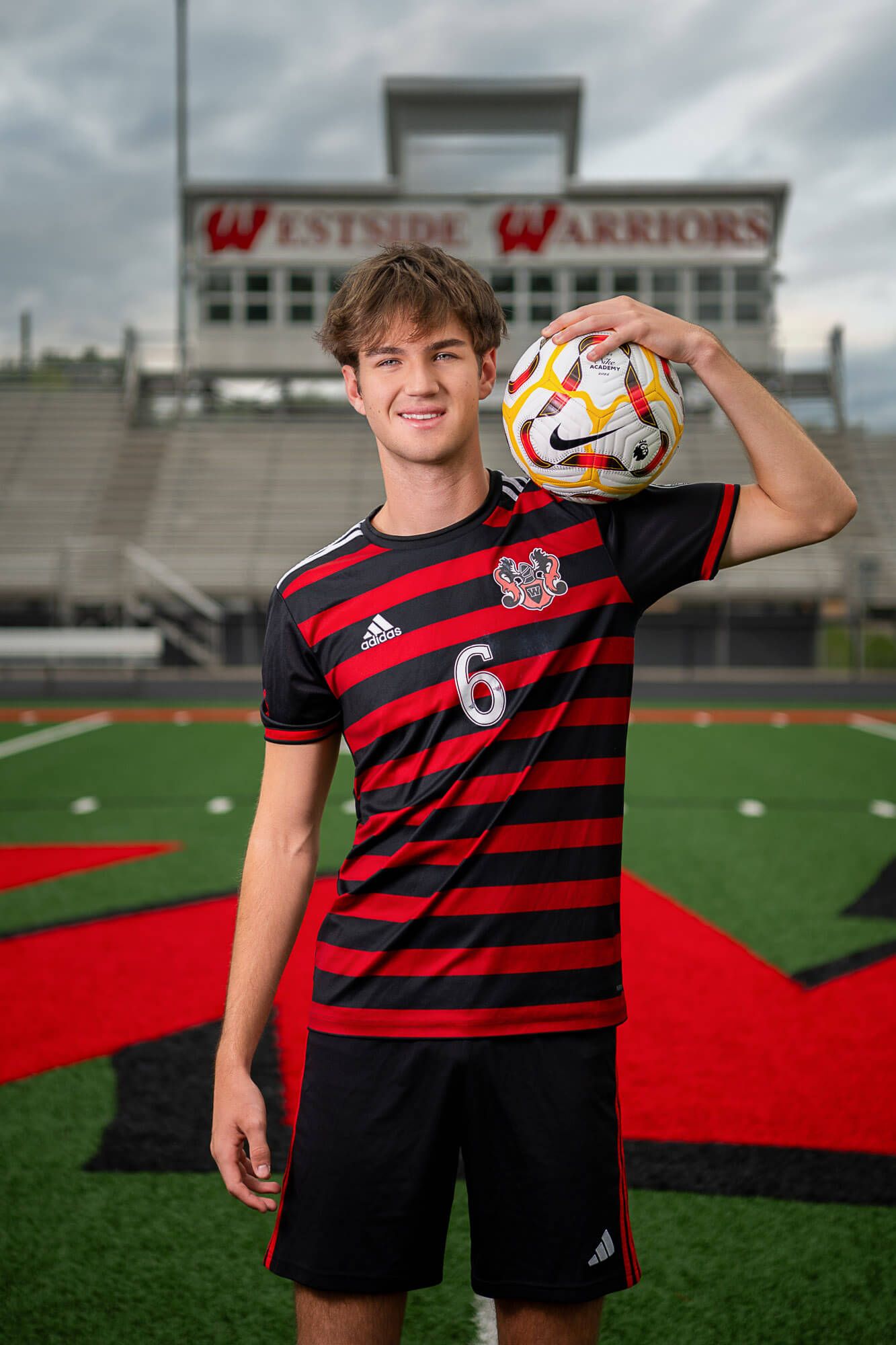 High school athlete poses in red and black striped jersey on soccer field with stadium backdrop.