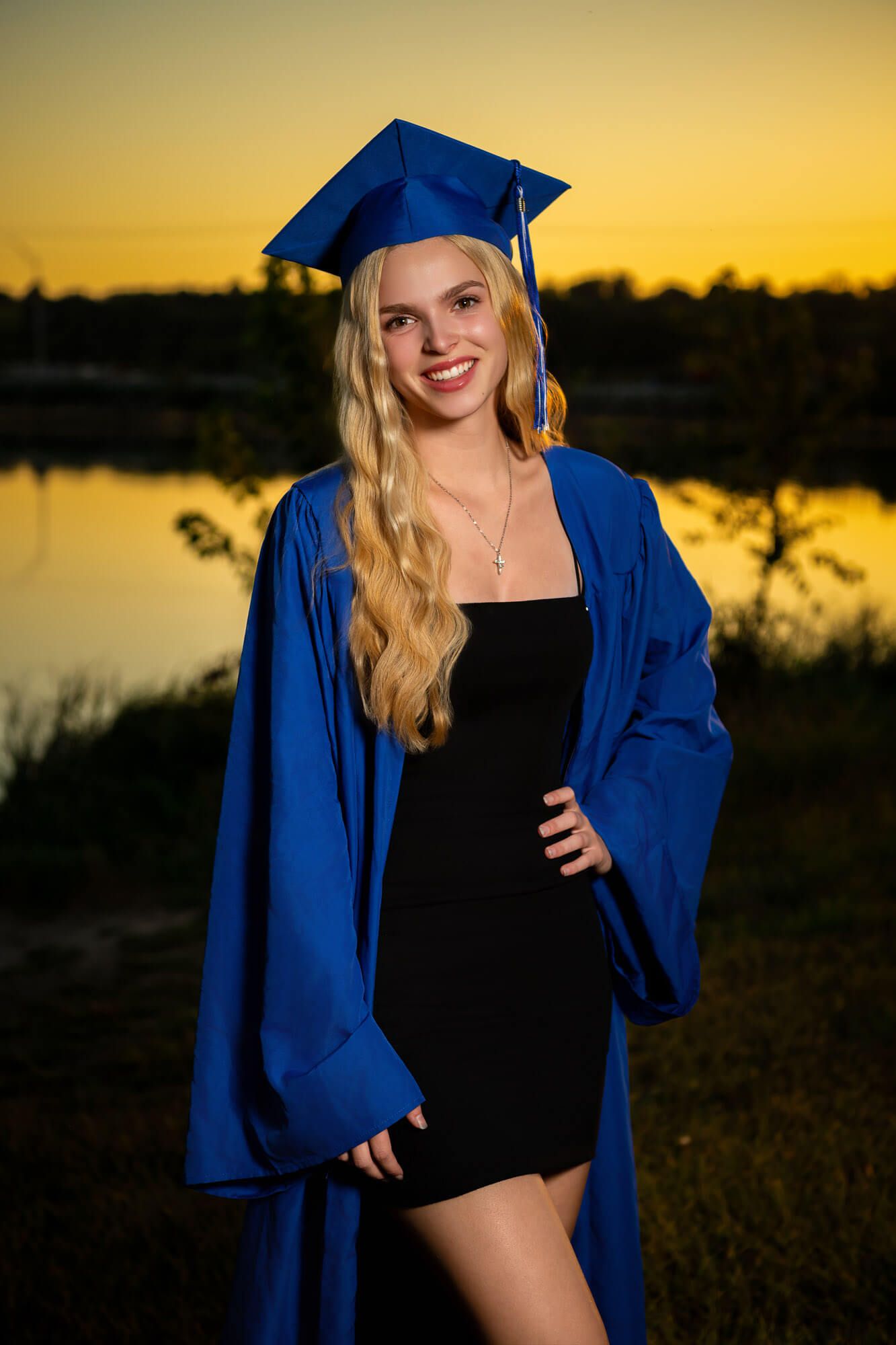 Graduation photos at sunset by a lake with blue cap and gown against golden sky.