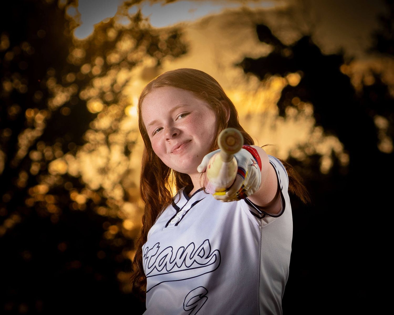 Young girl in softball uniform pointing a ball at camera with golden sunset bokeh background.