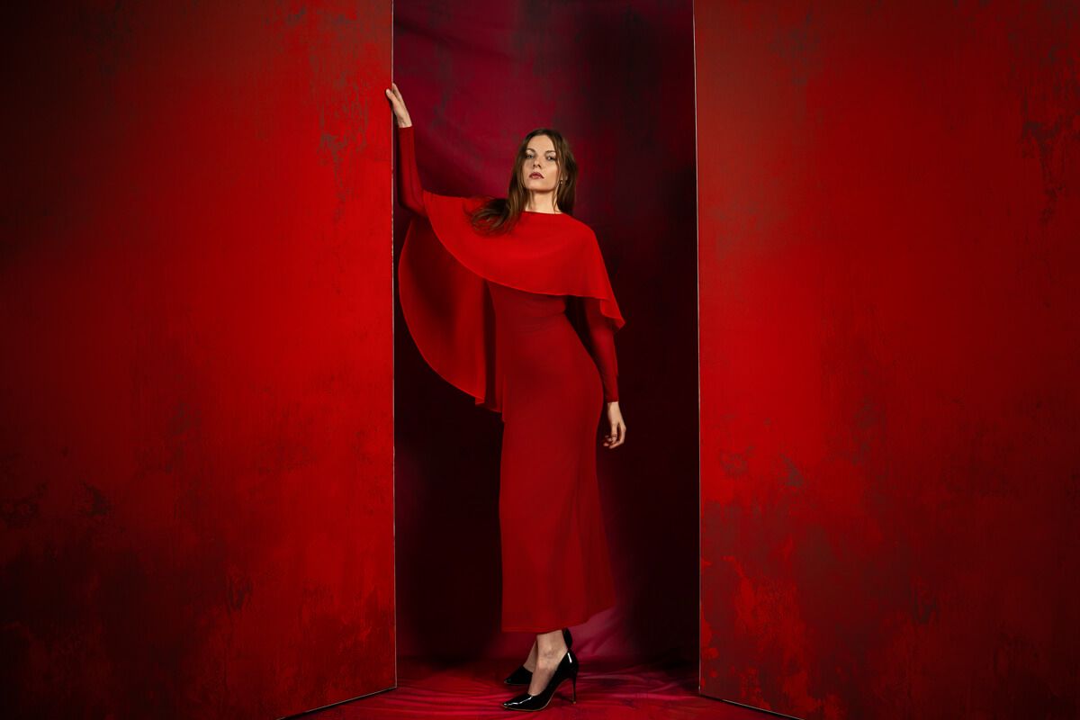 Woman in elegant red cape gown poses dramatically against bold red backdrop in striking fashion editorial.