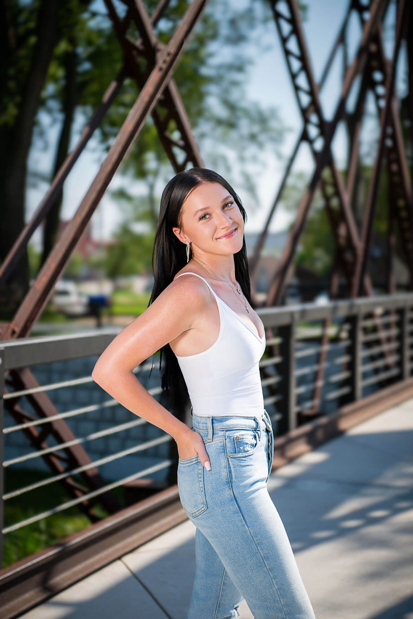 A person in a white tank top and light jeans poses against a metal bridge structure on a sunny day.