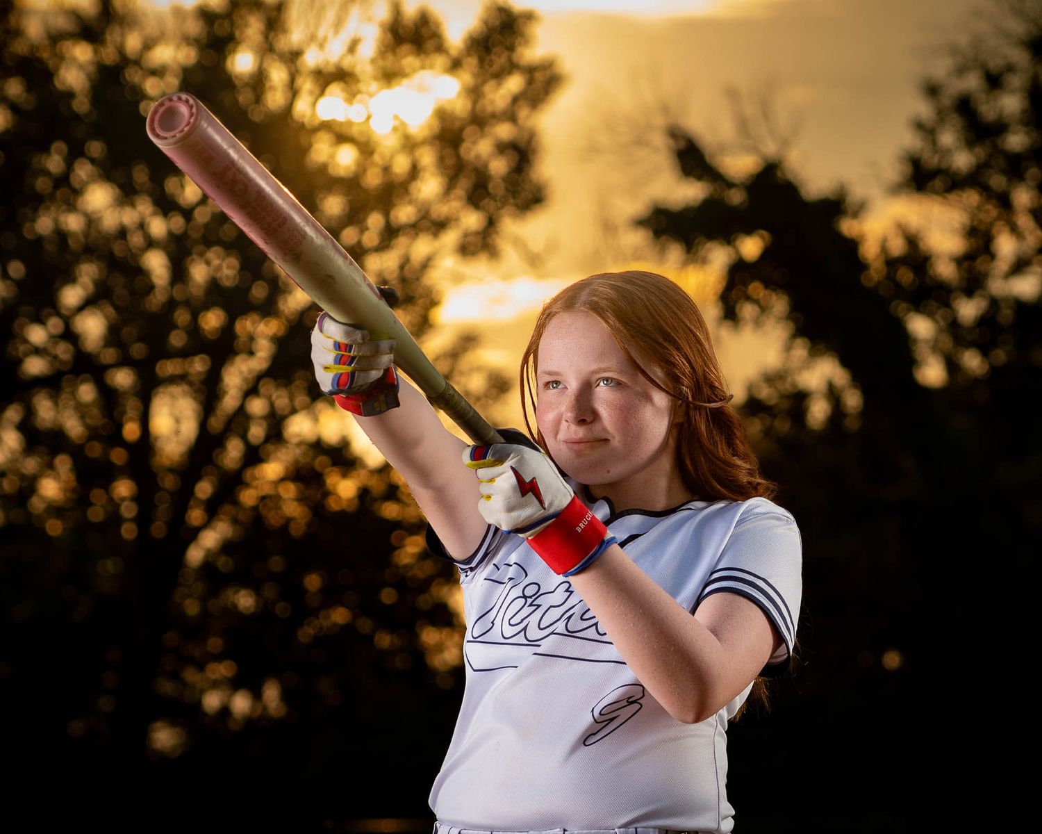 Young female softball player in uniform swings a bat at sunset with golden sky and trees in background.