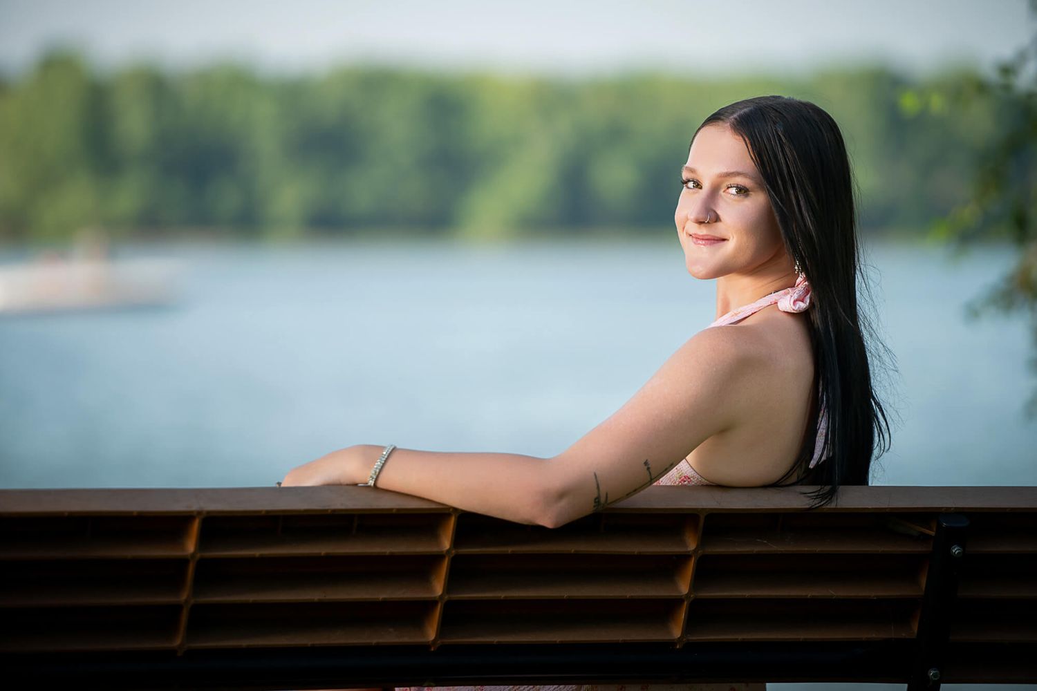Young woman with dark hair smiling over a wooden railing with a scenic lake and trees in the background.