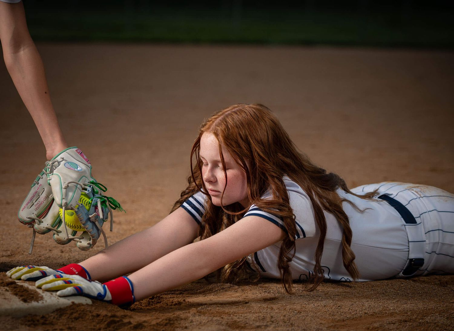 Young girl with red hair slides into base during softball game, reaching forward while player stands nearby.