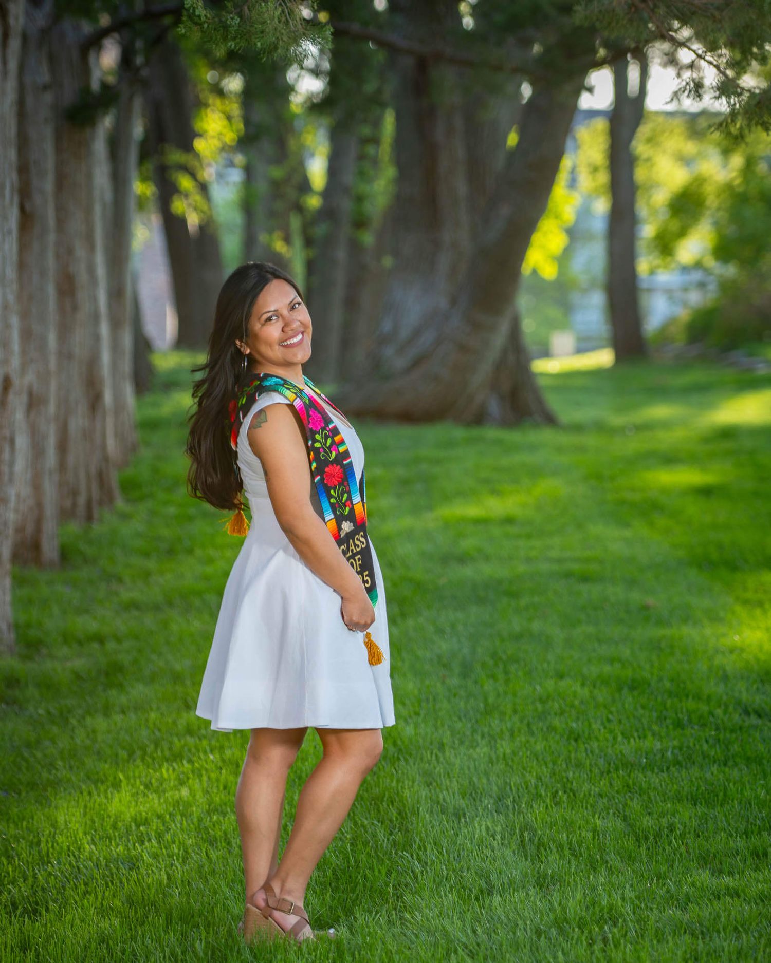 A smiling young woman in a white dress with colorful embroidered sash stands on green grass among tall trees.