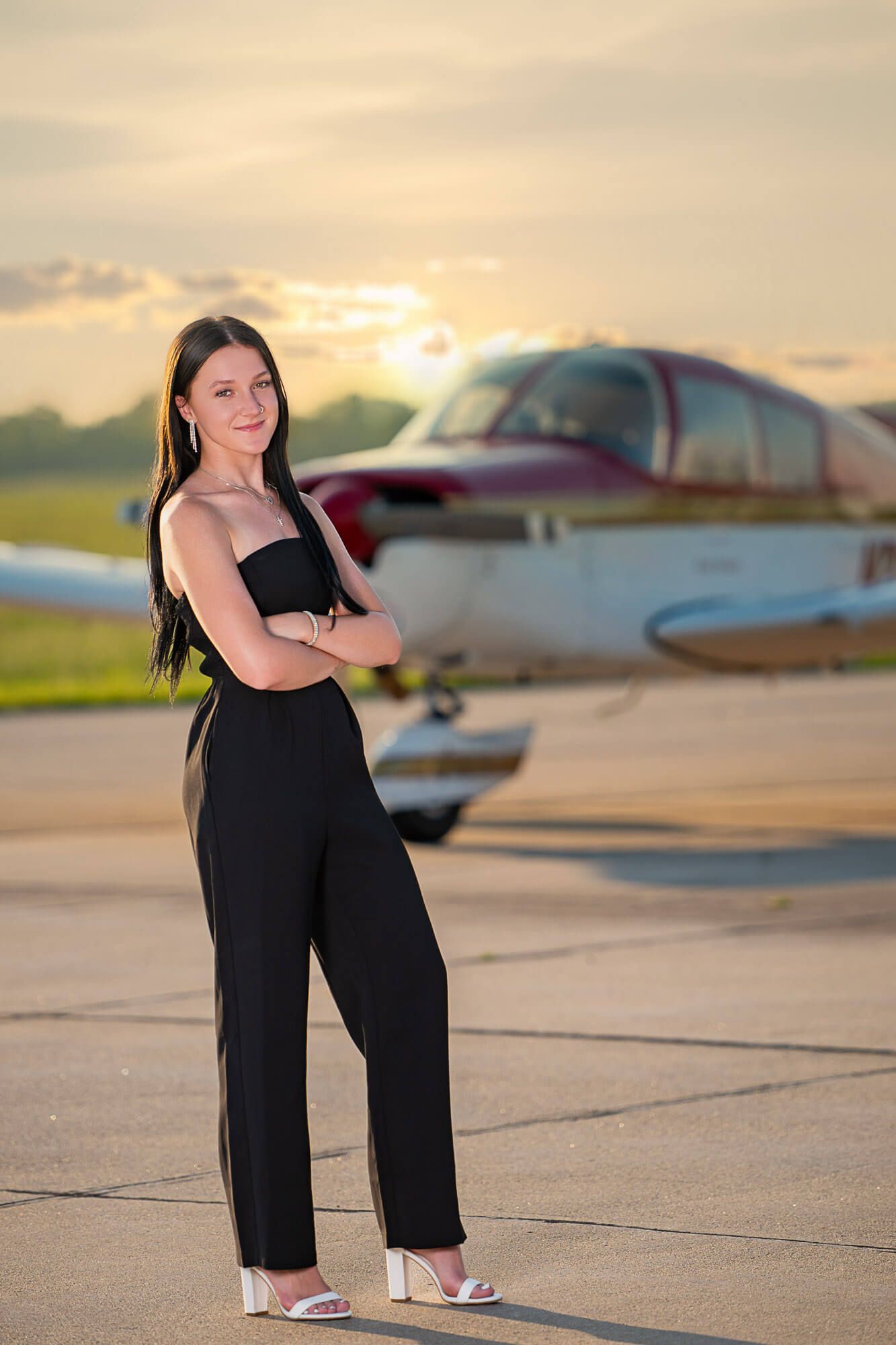 A figure in a black dress poses on an airport tarmac at sunset with small aircraft in the background.