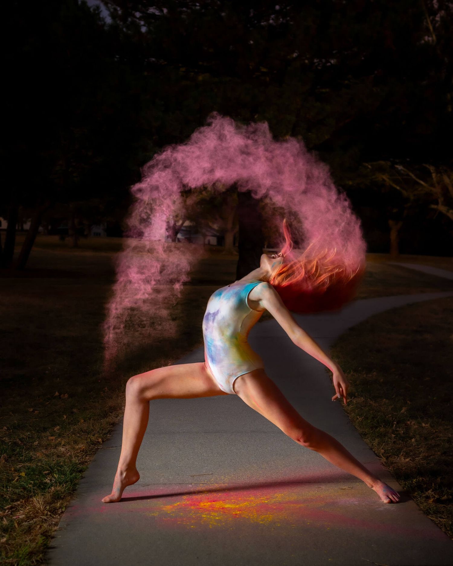 A dancer in white creates a cloud of pink powder while performing dynamic movements in a dark outdoor setting.