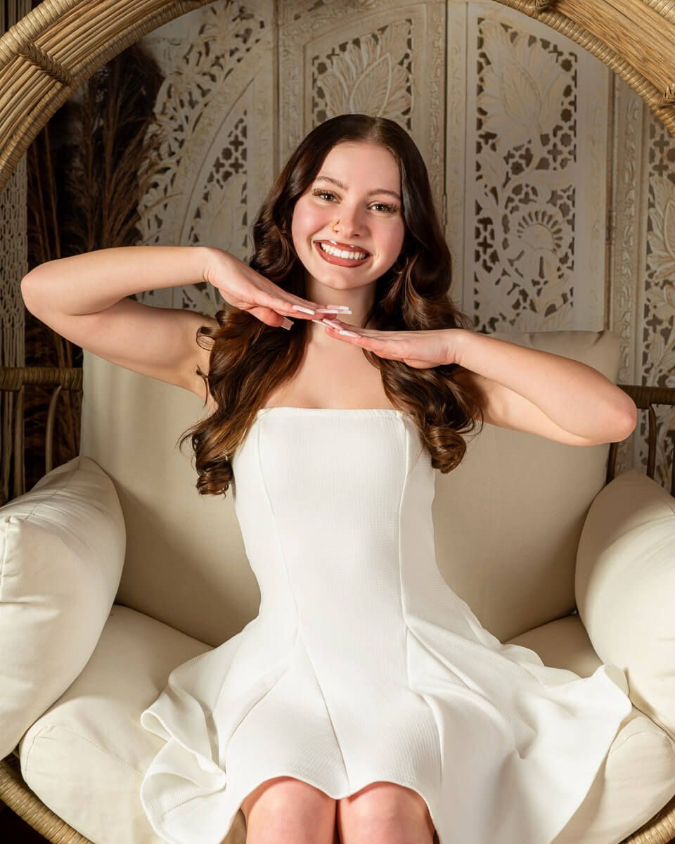 Young woman in white dress smiling while seated in a wicker chair with decorative backdrop.