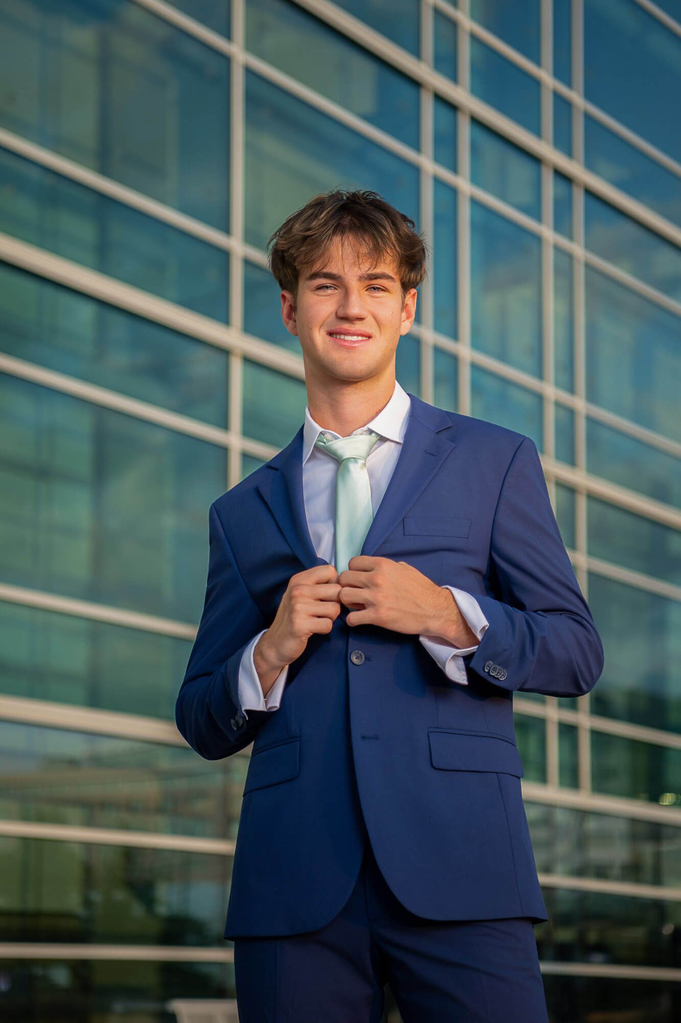 Omaha senior photography portrait of a Westside HS boy in a navy blue suit adjusting their tie against a modern glass building exterior.