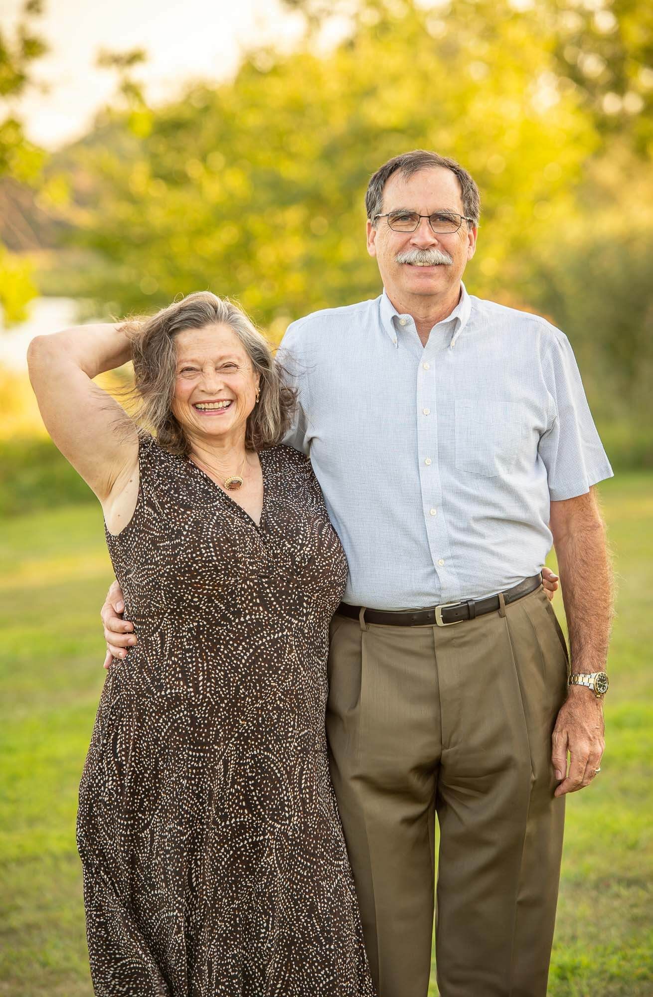 A mature couple poses together outdoors during golden hour with autumn foliage in the background.