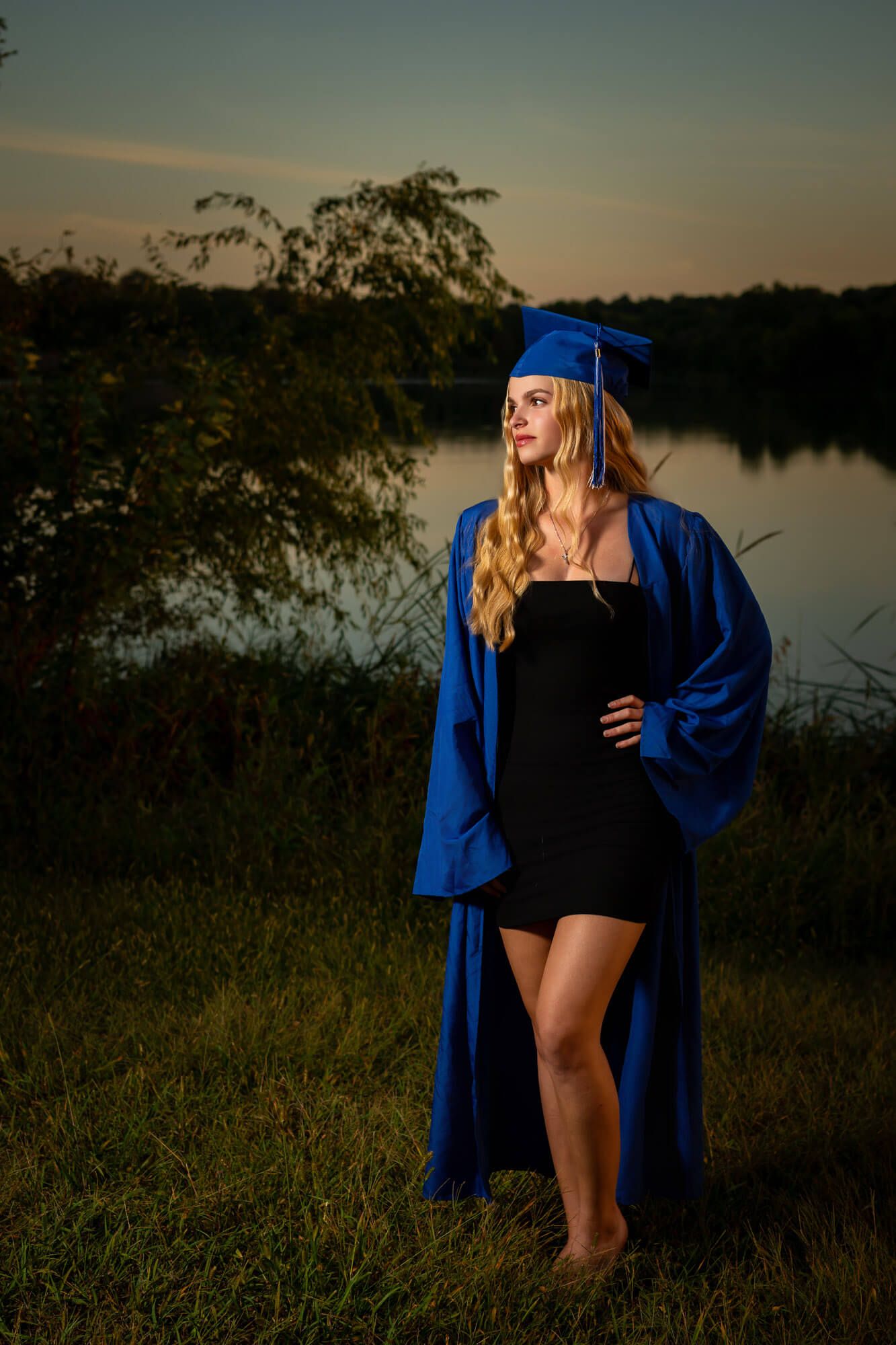 Blonde graduate in blue cap and gown posing by lake at dusk senior portrait.