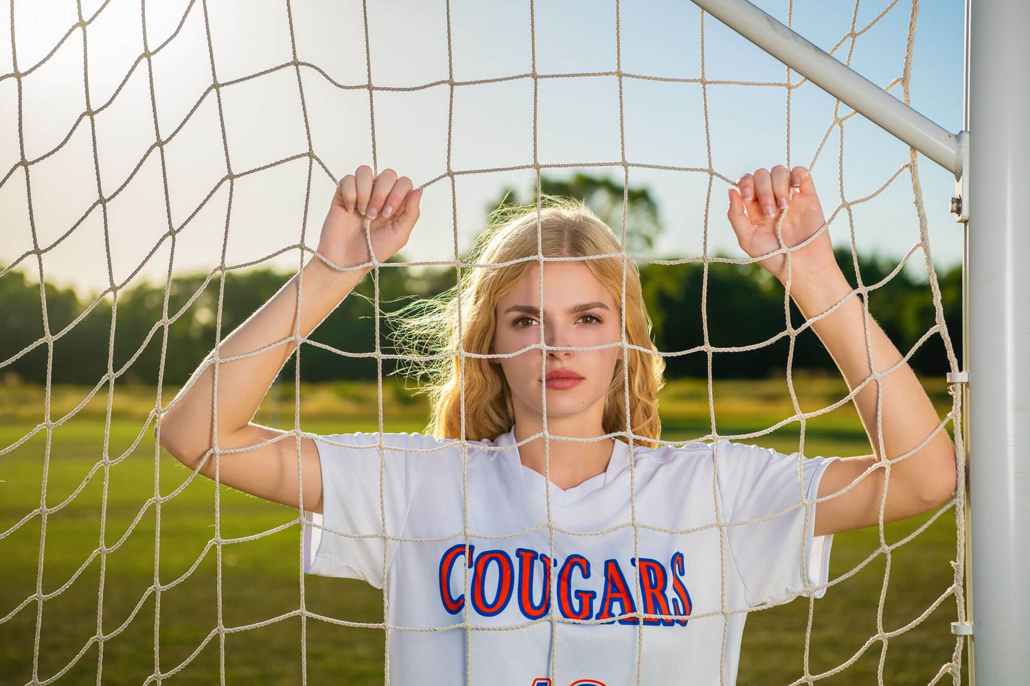 Blonde teen in Cougars jersey holding soccer goal net on green field at golden hour.
