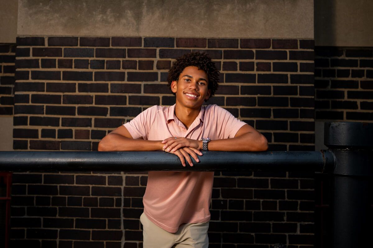 Smiling young man in pink polo leans on a blue railing against a brick wall in warm evening light.