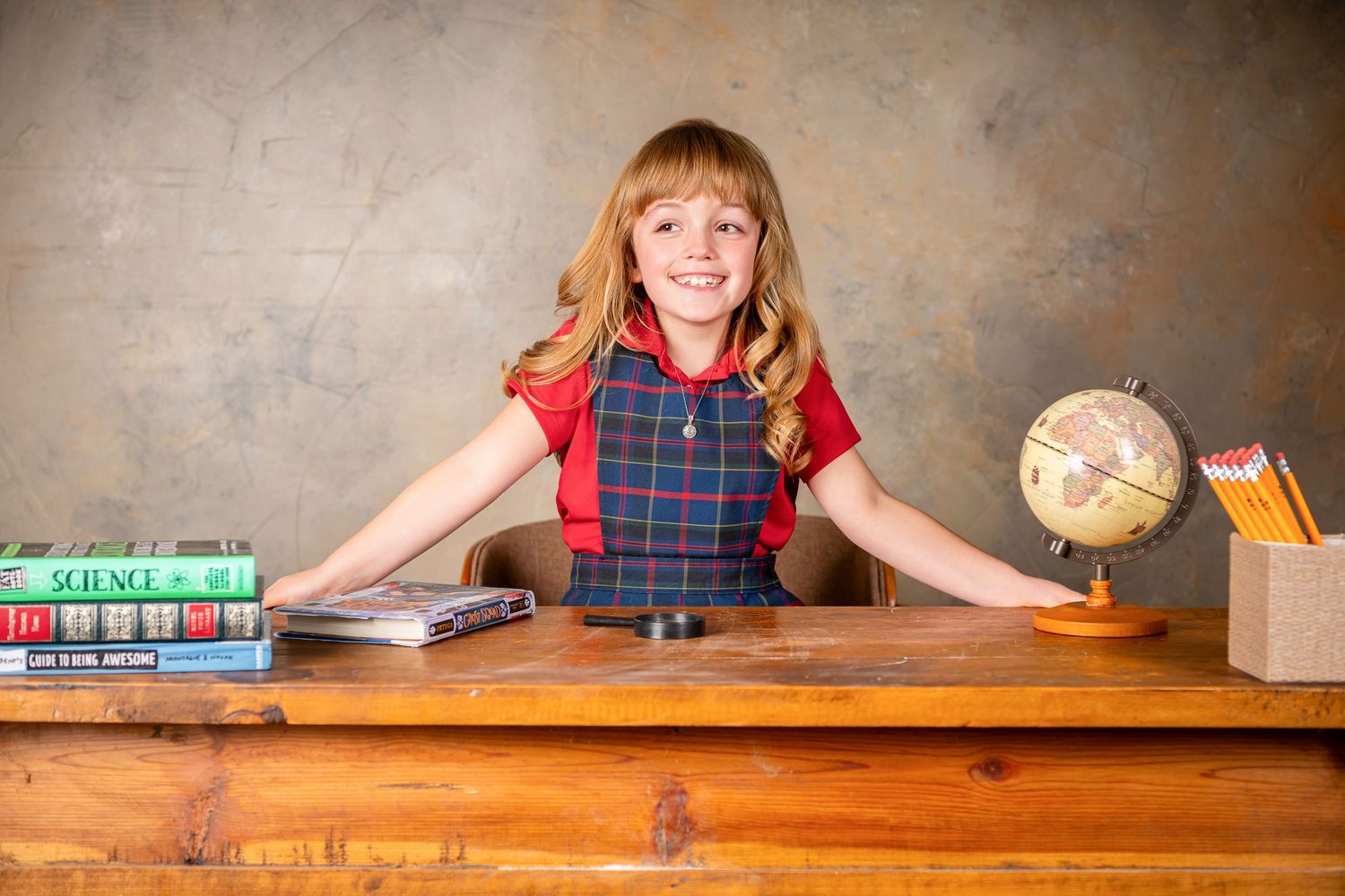 Student in plaid uniform sits at wooden desk with textbooks, globe and pencils in classroom setting.