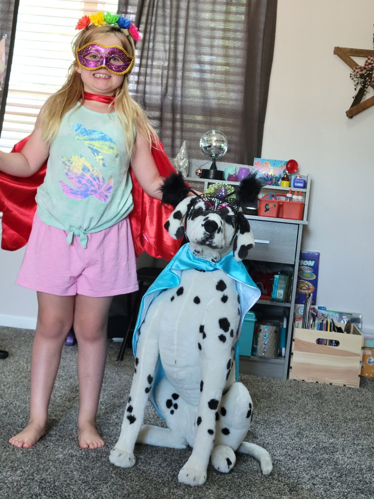 Dalmatian dog wearing blue outfit poses next to young person in colorful costume and rainbow accessories.