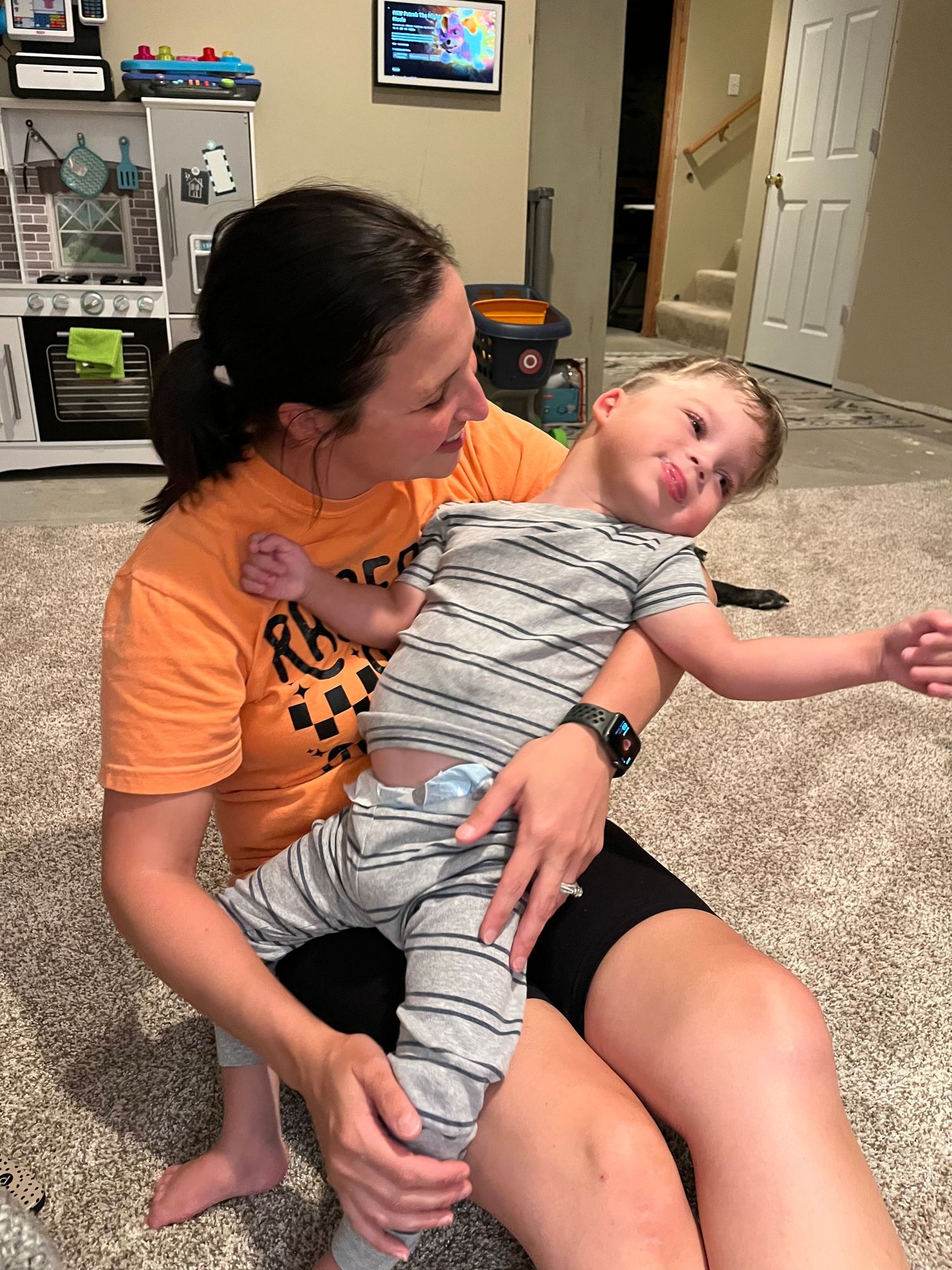 A person in an orange shirt holds a sleepy young child wearing a striped outfit while sitting on a carpeted floor.