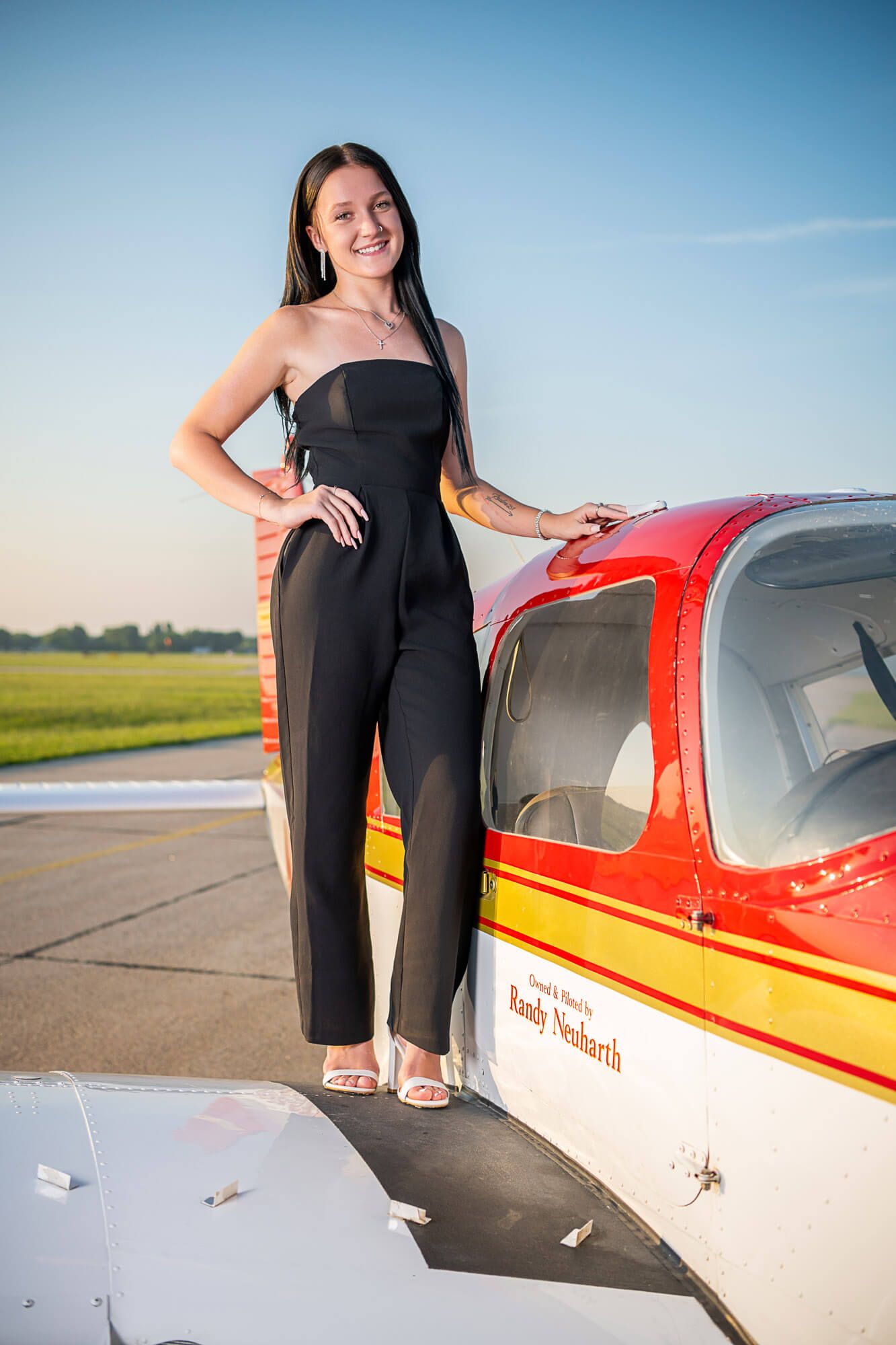 A person in a black jumpsuit poses next to a red and white small aircraft on an airfield runway.