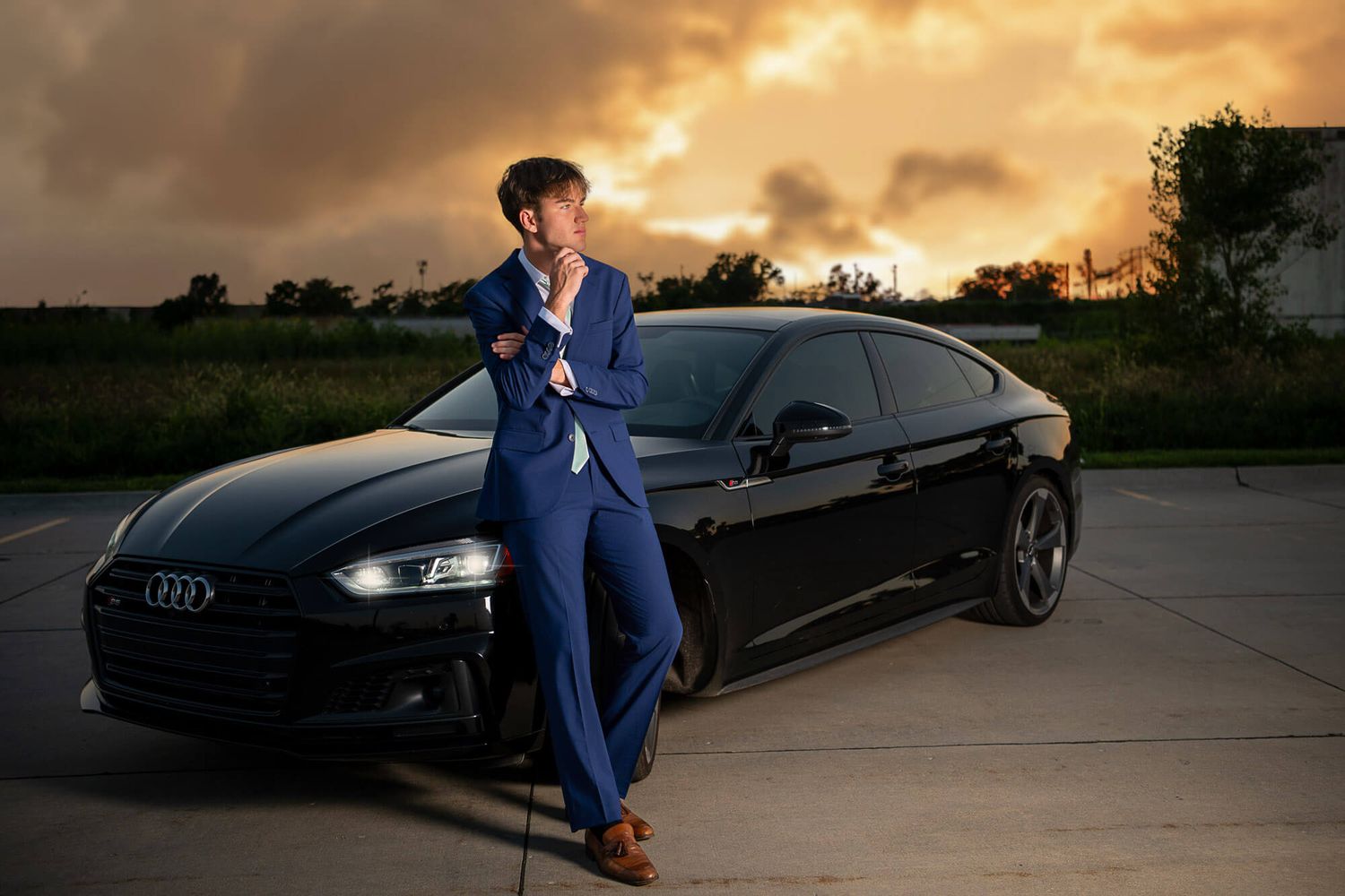 A person in a blue suit poses with a black Audi car during golden hour sunset.