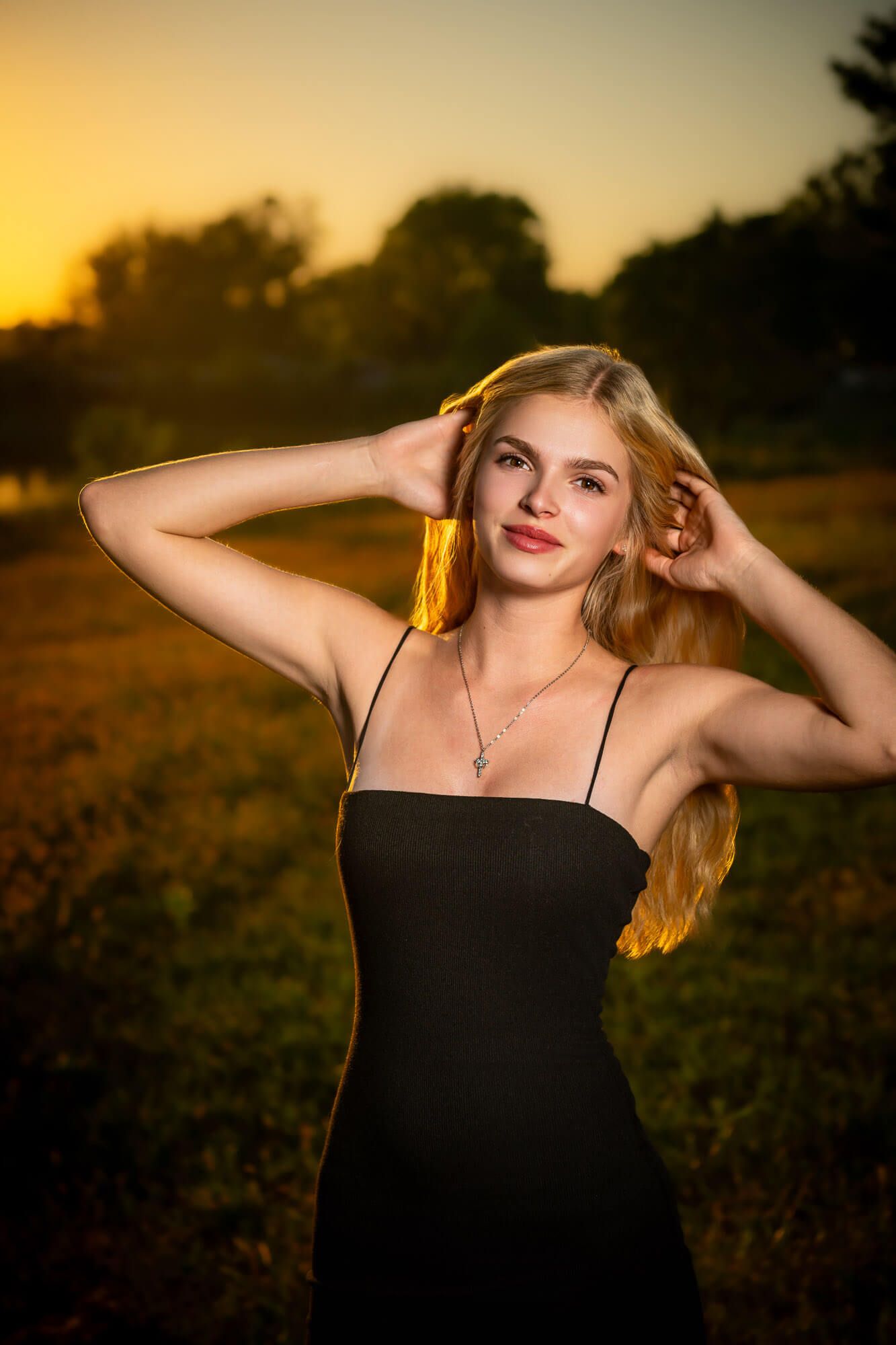 A high school senior from Gross Catholic in a black dress poses gracefully against a golden sunset backdrop in a natural outdoor setting.