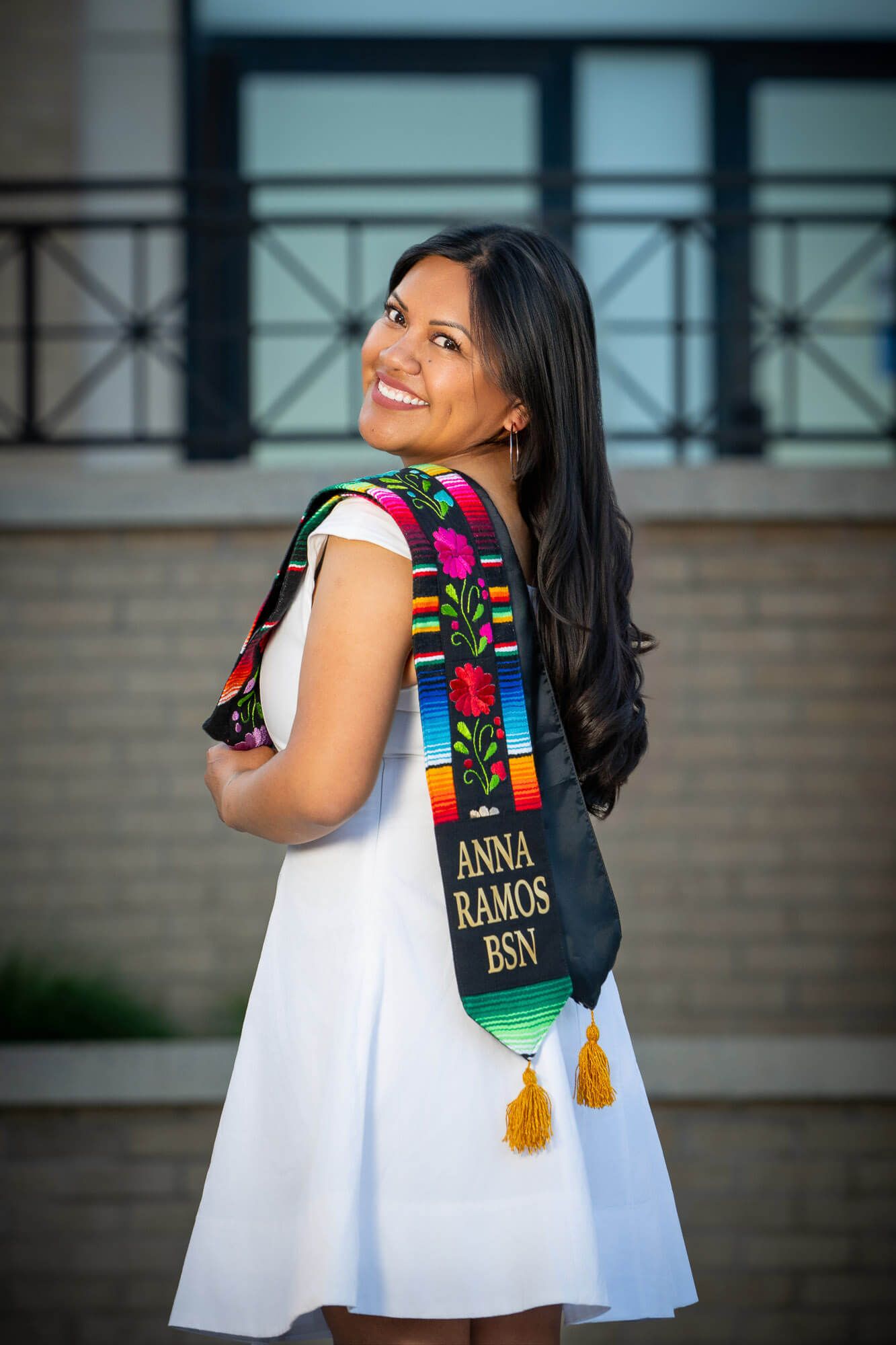 Anna Ramos smiles in white dress wearing colorful serape sash and BSN graduation stole outdoors.