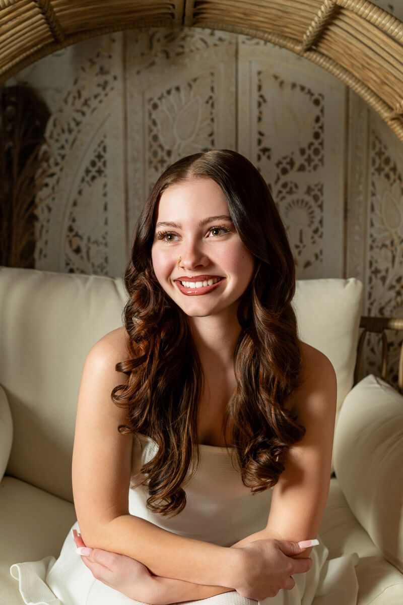 Smiling young woman with long curly brown hair wearing a white dress, seated on a cream sofa with decorative backdrop.