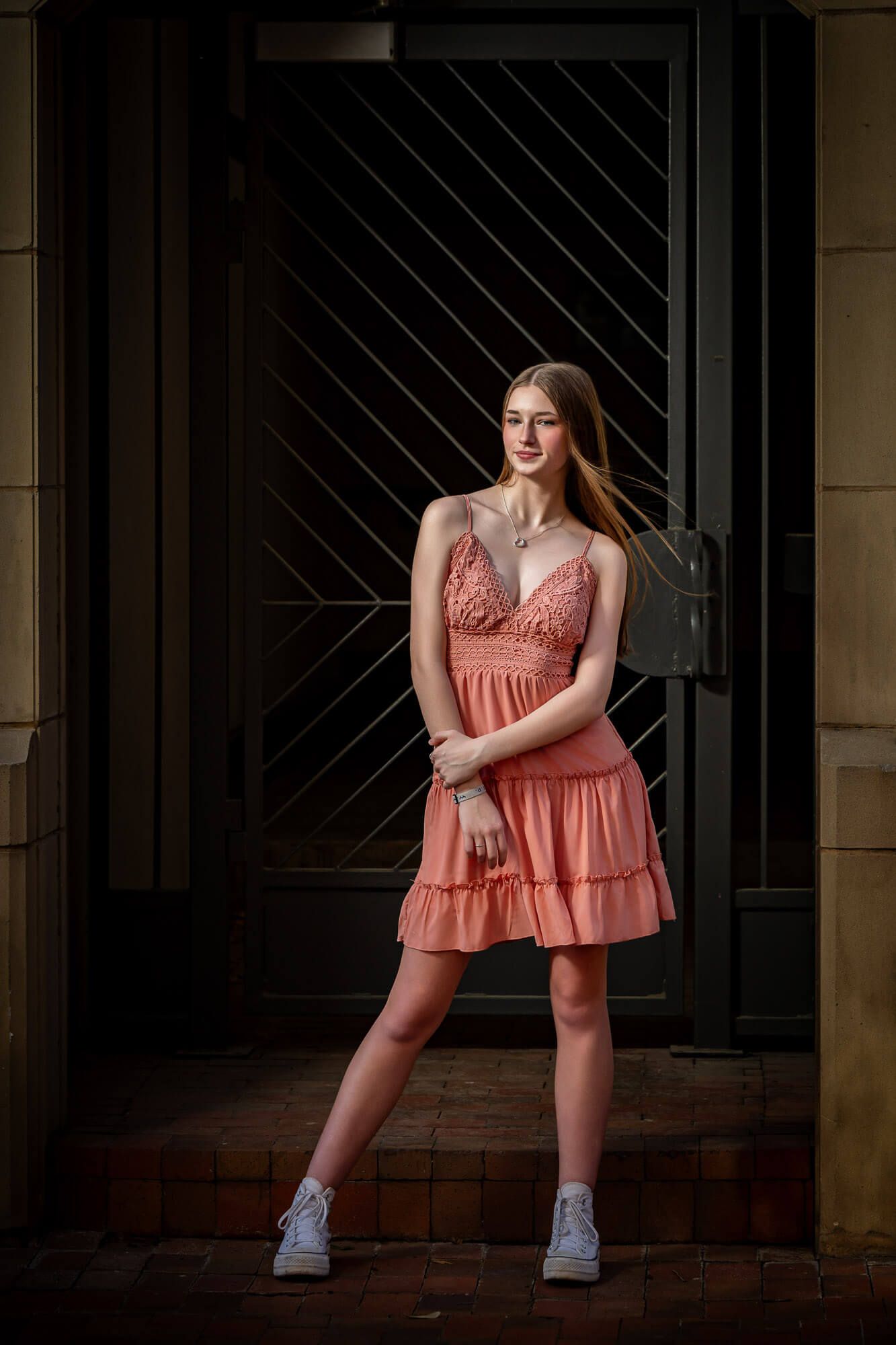 Young woman in coral mini dress and white sneakers posing elegantly in front of dark ornate metal doors.