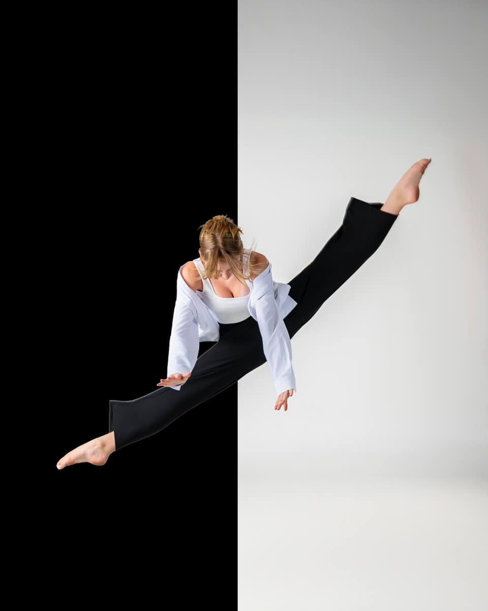 A dancer leaps mid-air in a split jump, wearing white top and black pants against a bold black and white background.