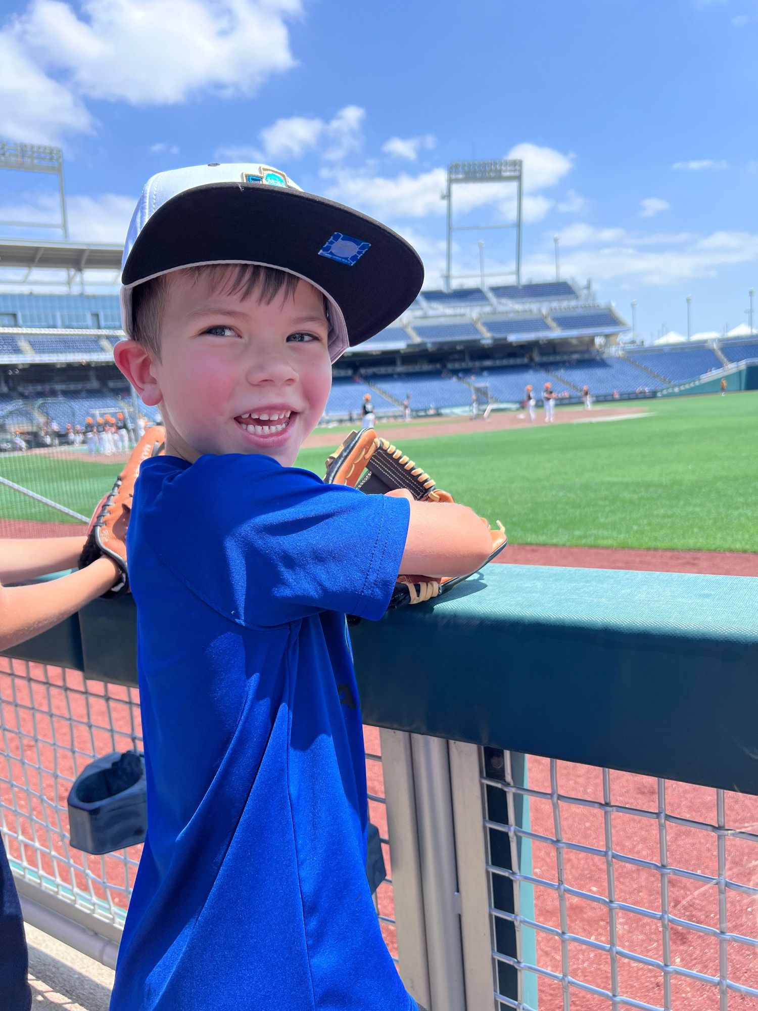 Young baseball fan in a blue shirt and cap enjoys a sunny day at a baseball stadium with empty seats visible in background.