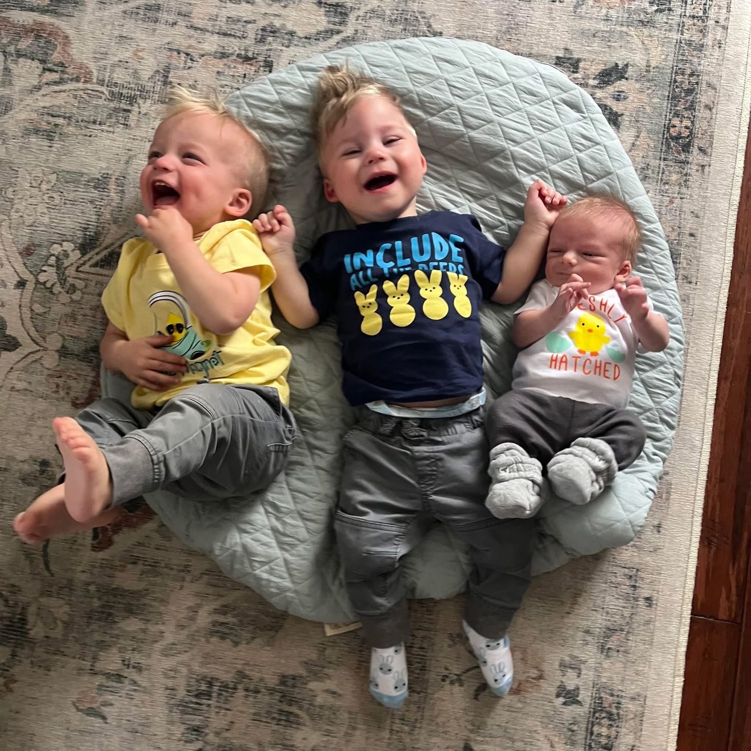 Three laughing babies lying together on a light gray cushioned surface while wearing casual outfits.