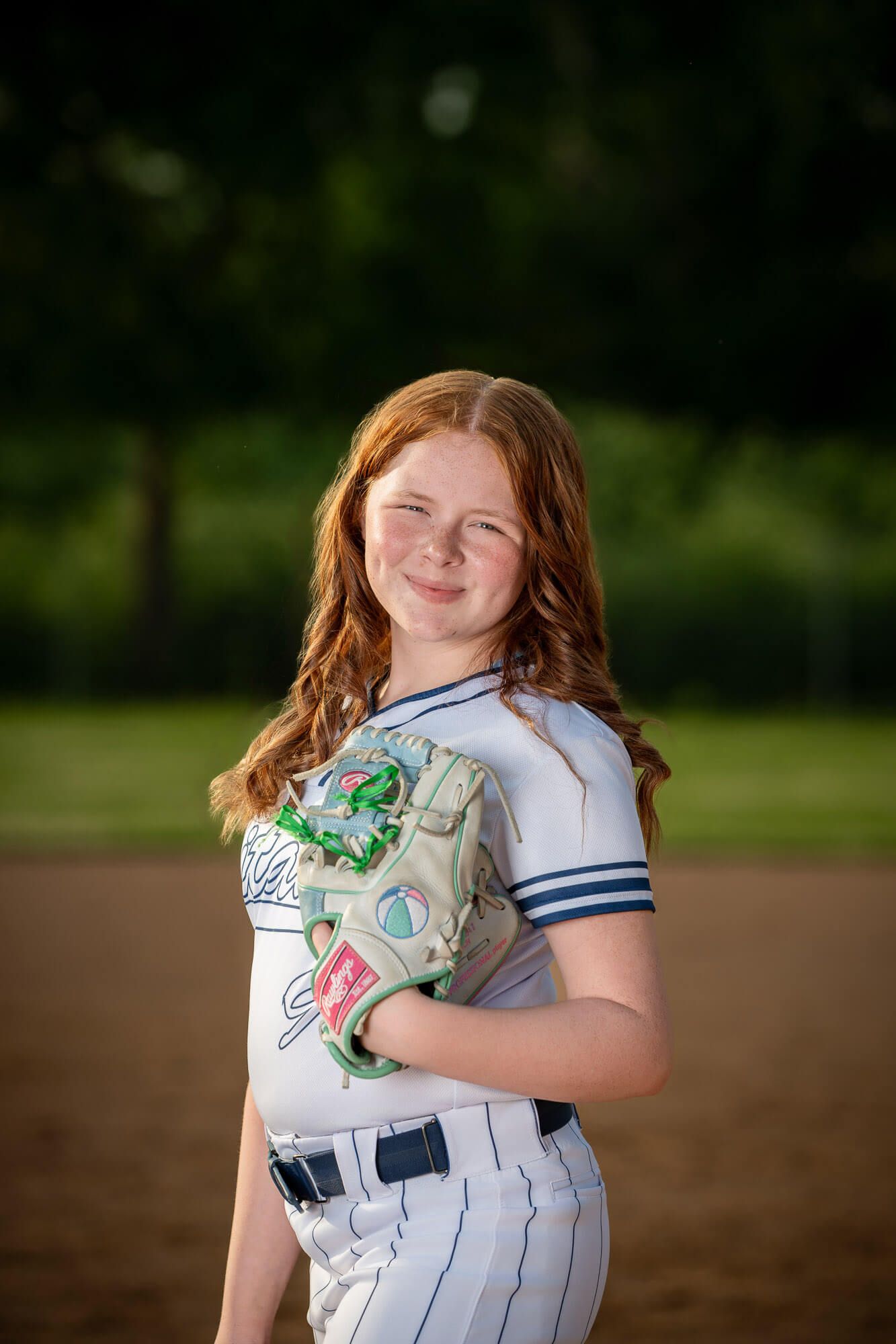 Young redhead girl in softball uniform holding a colorful glove, posing on a dirt field with green trees behind.