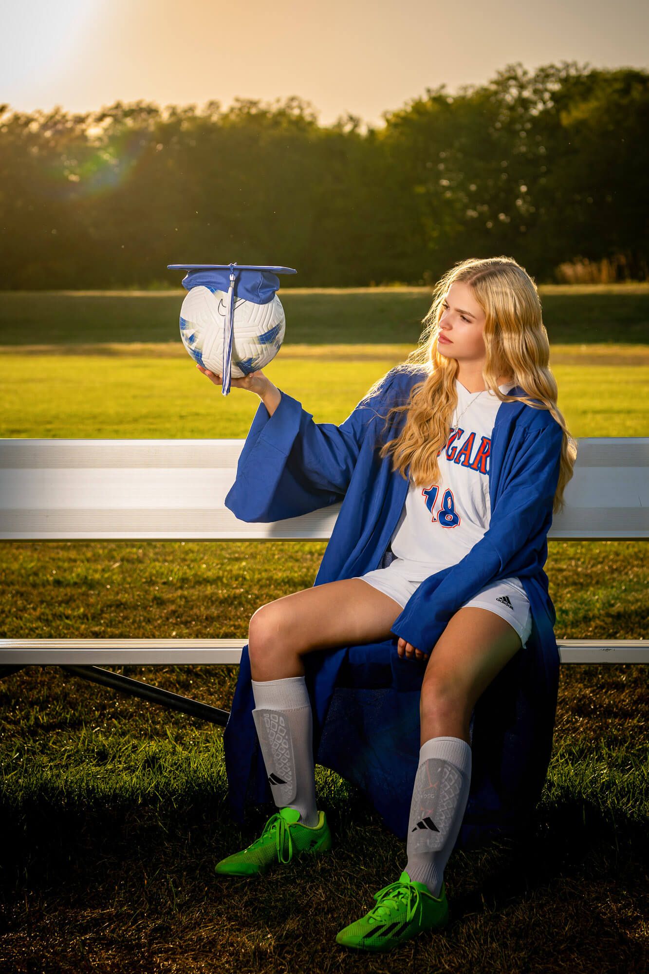 Female soccer player in blue graduation gown holding cap and soccer ball while sitting on bench at sunset.