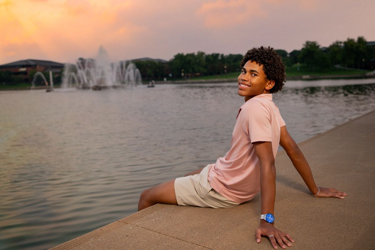 Young Black boy sitting by a lakeside fountain at sunset, wearing a pink shirt and white shorts, smiling over his shoulder.