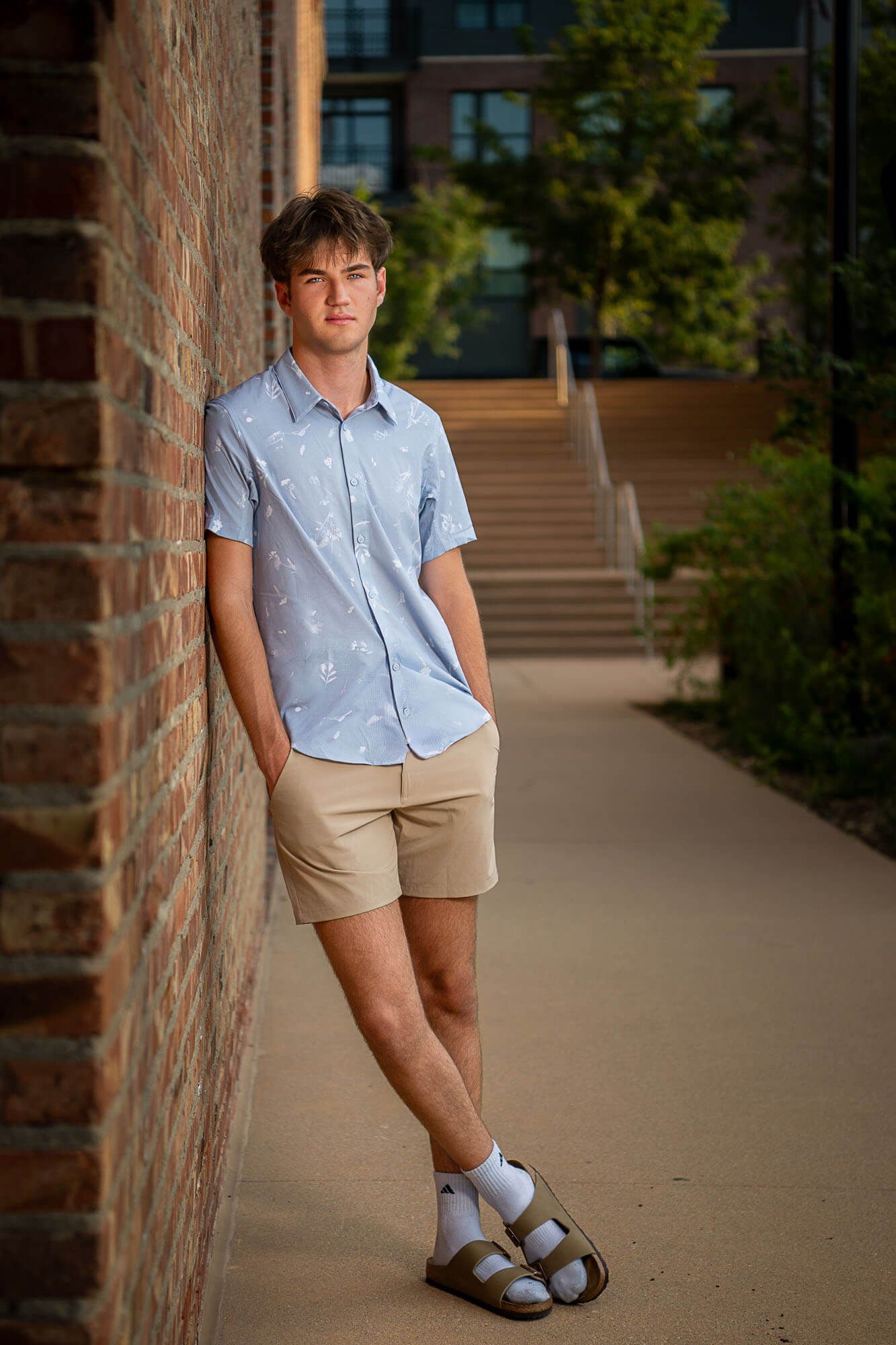 A person in a light blue shirt and khaki shorts poses casually against a brick wall in an outdoor walkway.