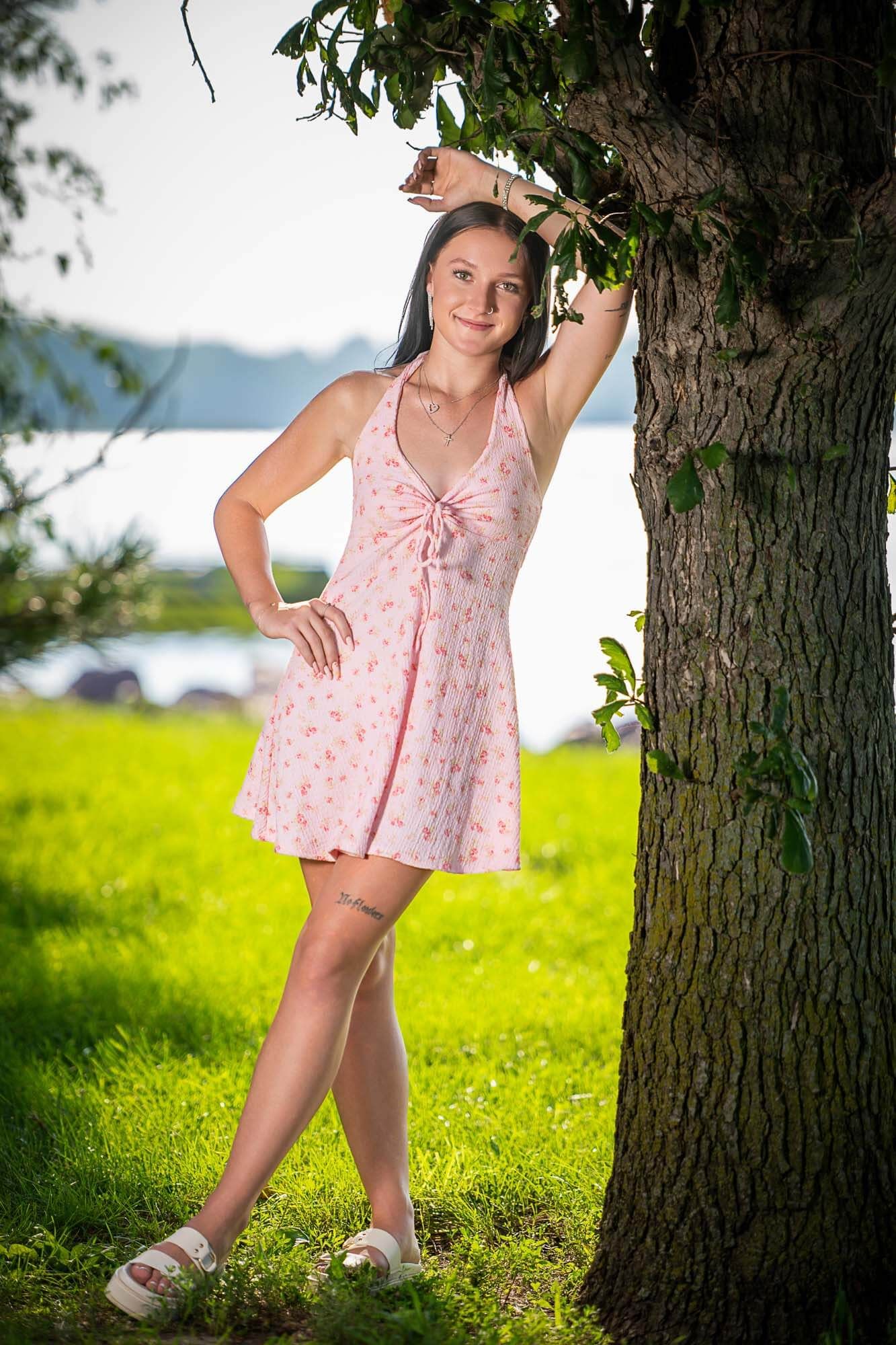 A pink floral sundress and sandals are worn on a sunny day by a tree with water and mountains in the background.