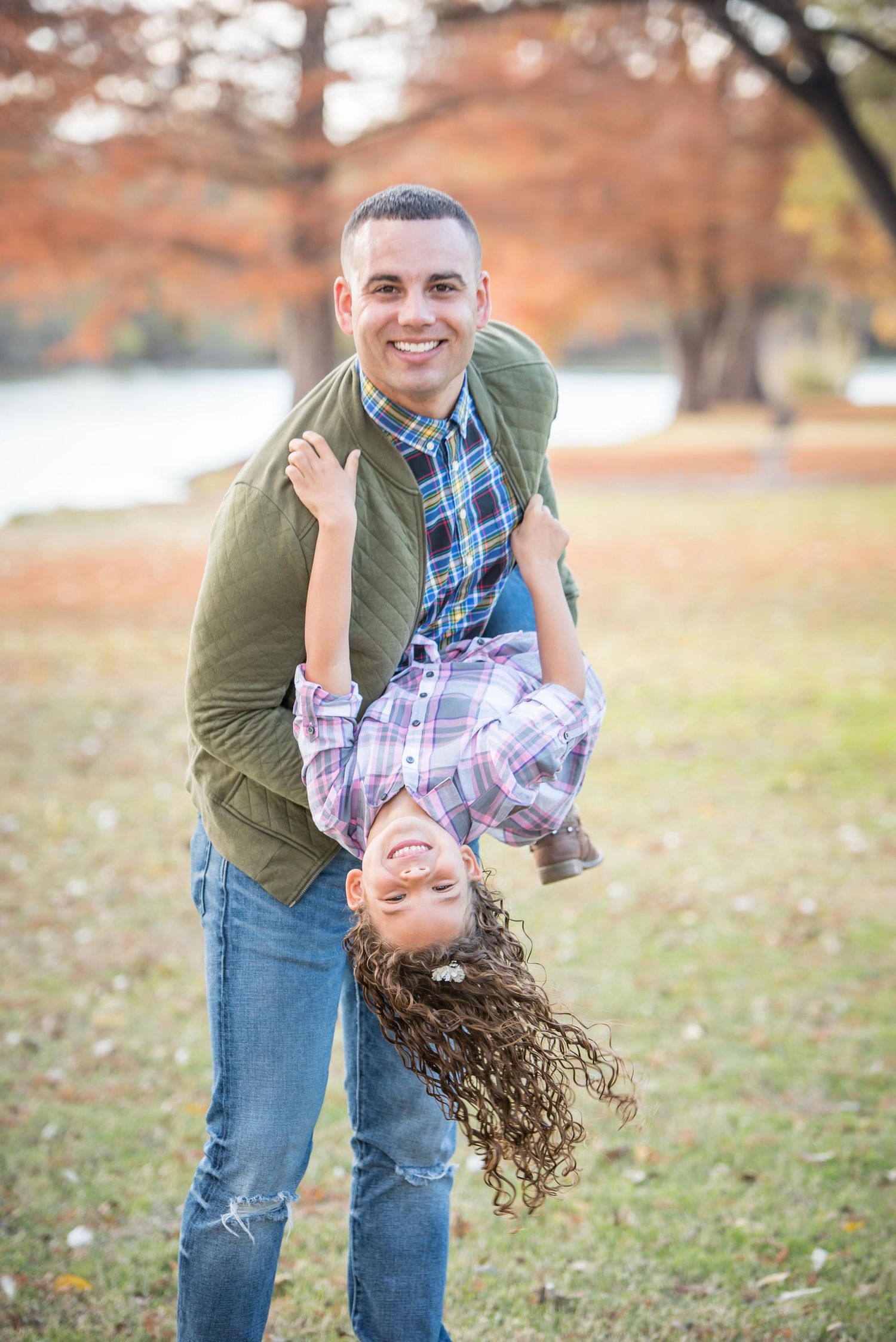 Dad in green jacket playfully holds daughter upside down while both smile in an autumn park setting for Omaha family photography session.