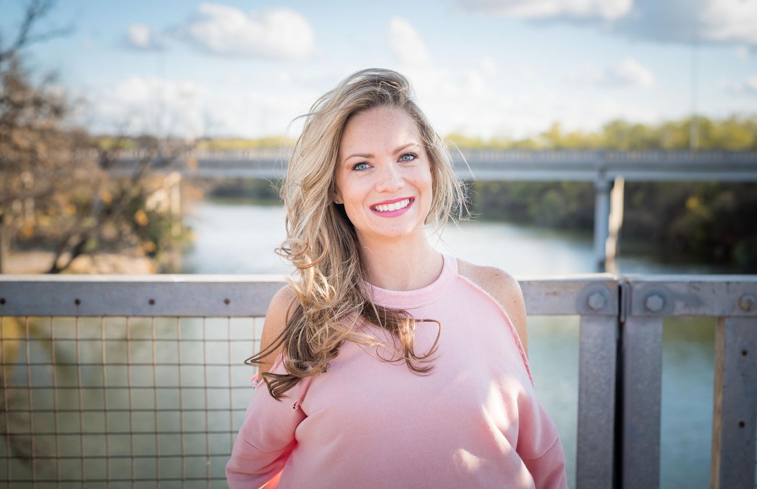 A person in a pink sweater smiles while standing on a bridge over water on a sunny day.