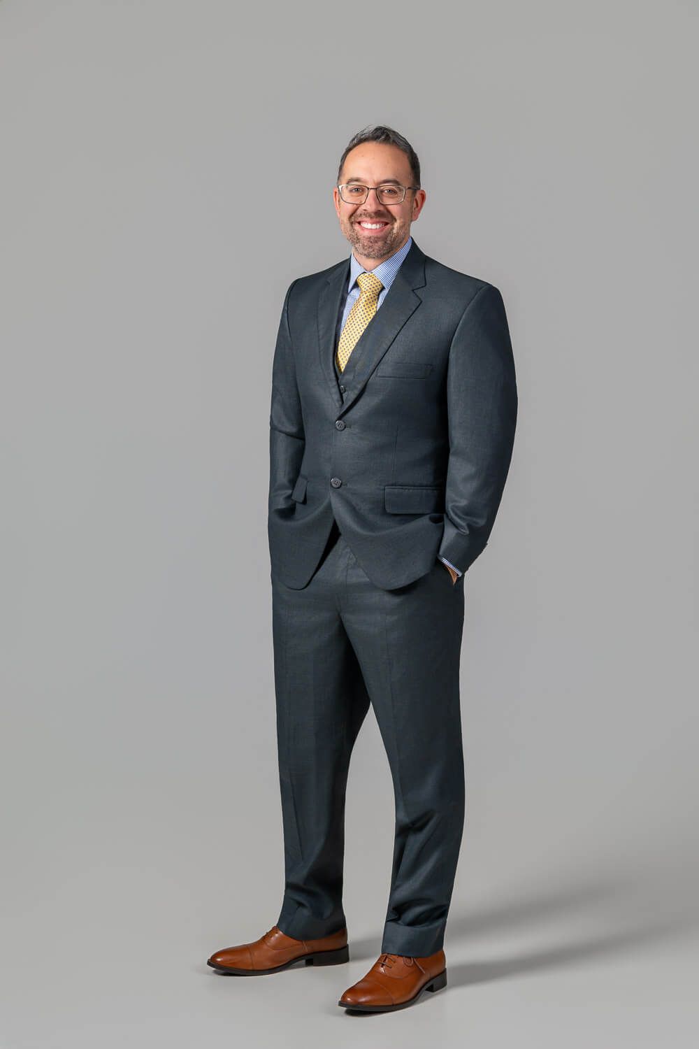 Professional business portrait of someone wearing a dark gray suit with yellow tie and brown shoes against gray background.