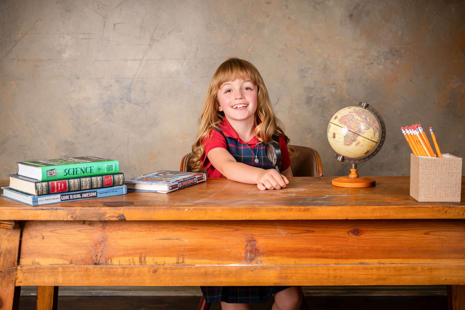 Student sits at wooden desk with textbooks, globe and pencil holder in classroom setting.