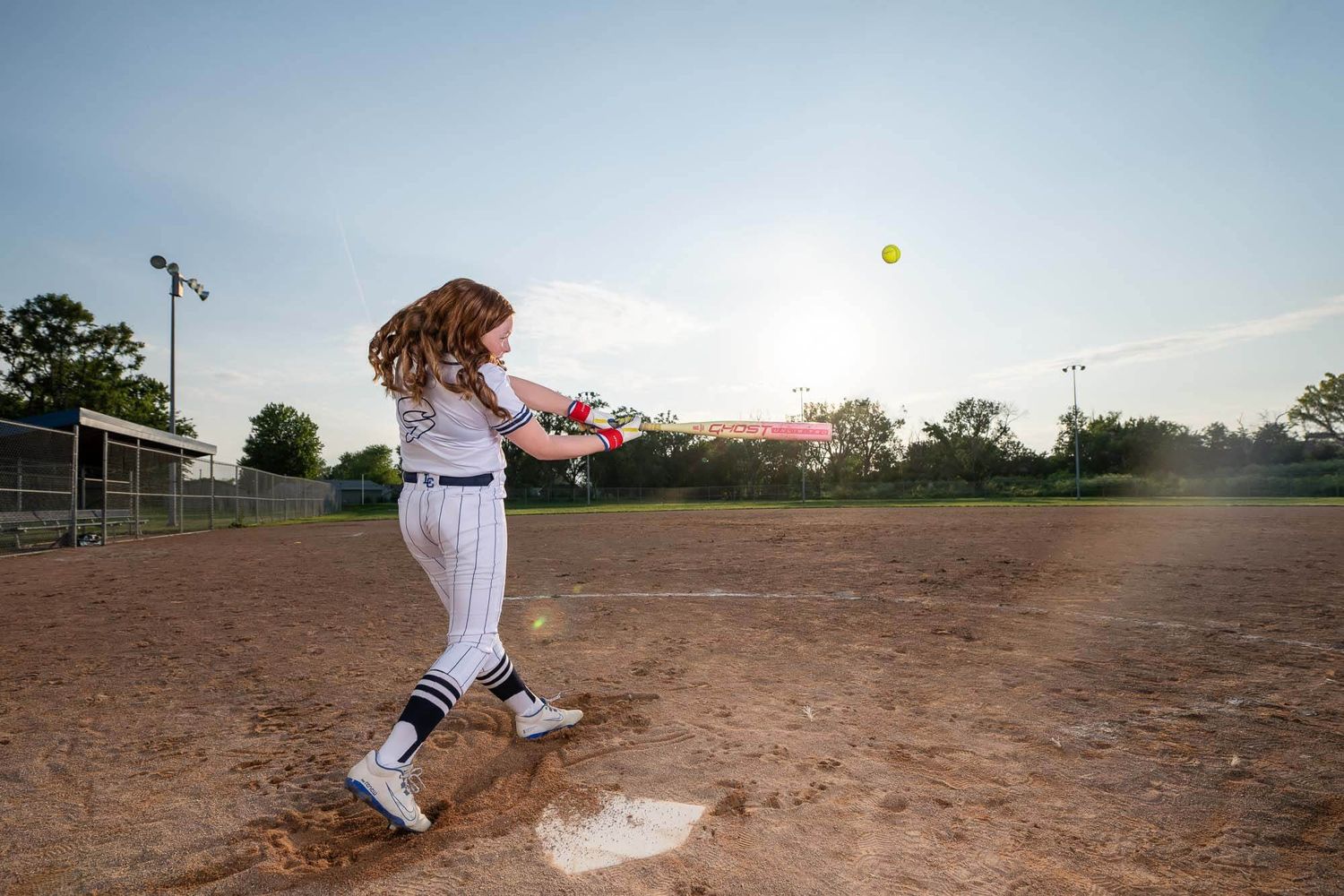 A young girl in a white uniform swings a bat at a softball during a game on a dirt field at sunset.