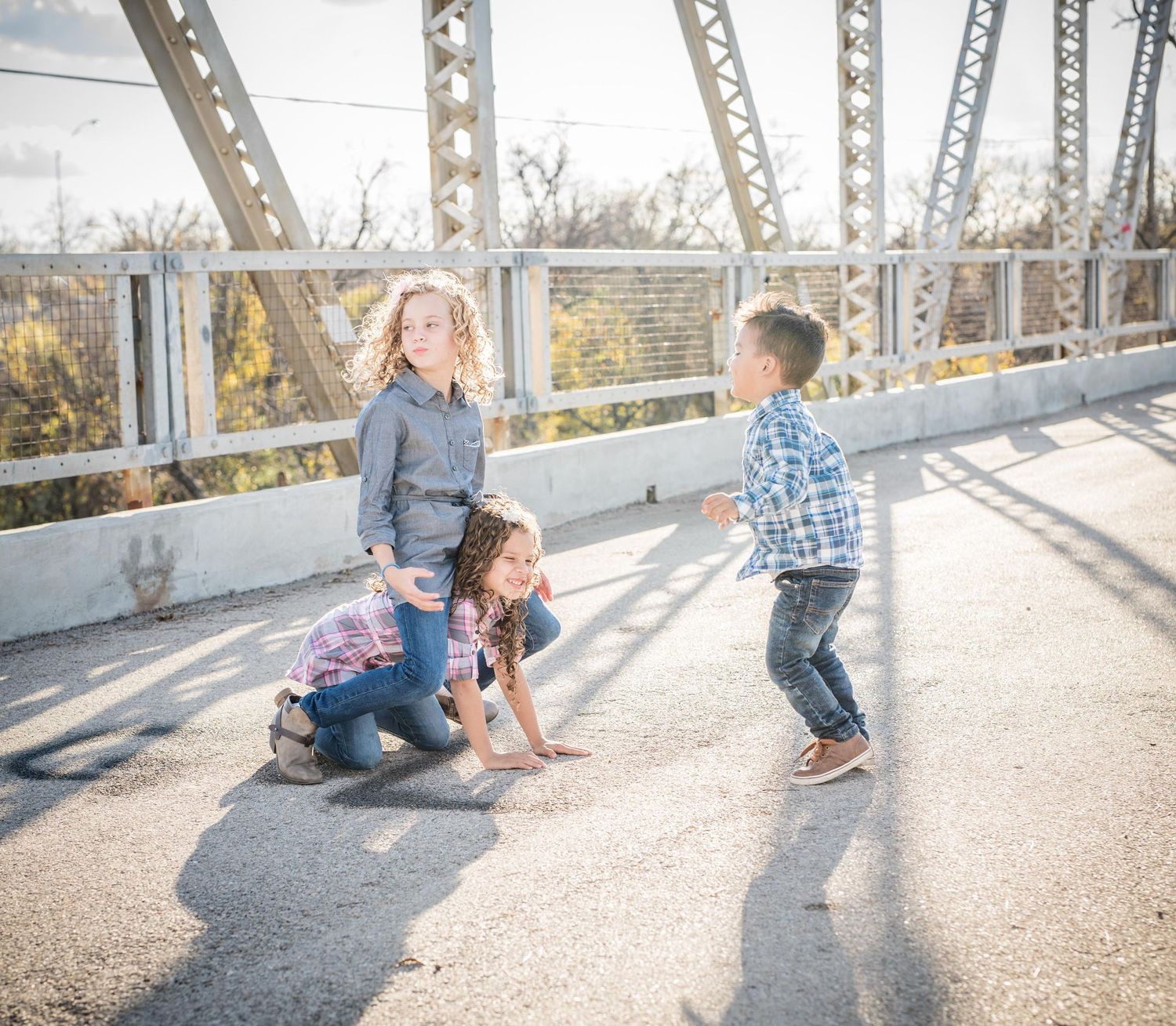 Three people playing and laughing together on a sunlit metal bridge on a bright autumn afternoon.