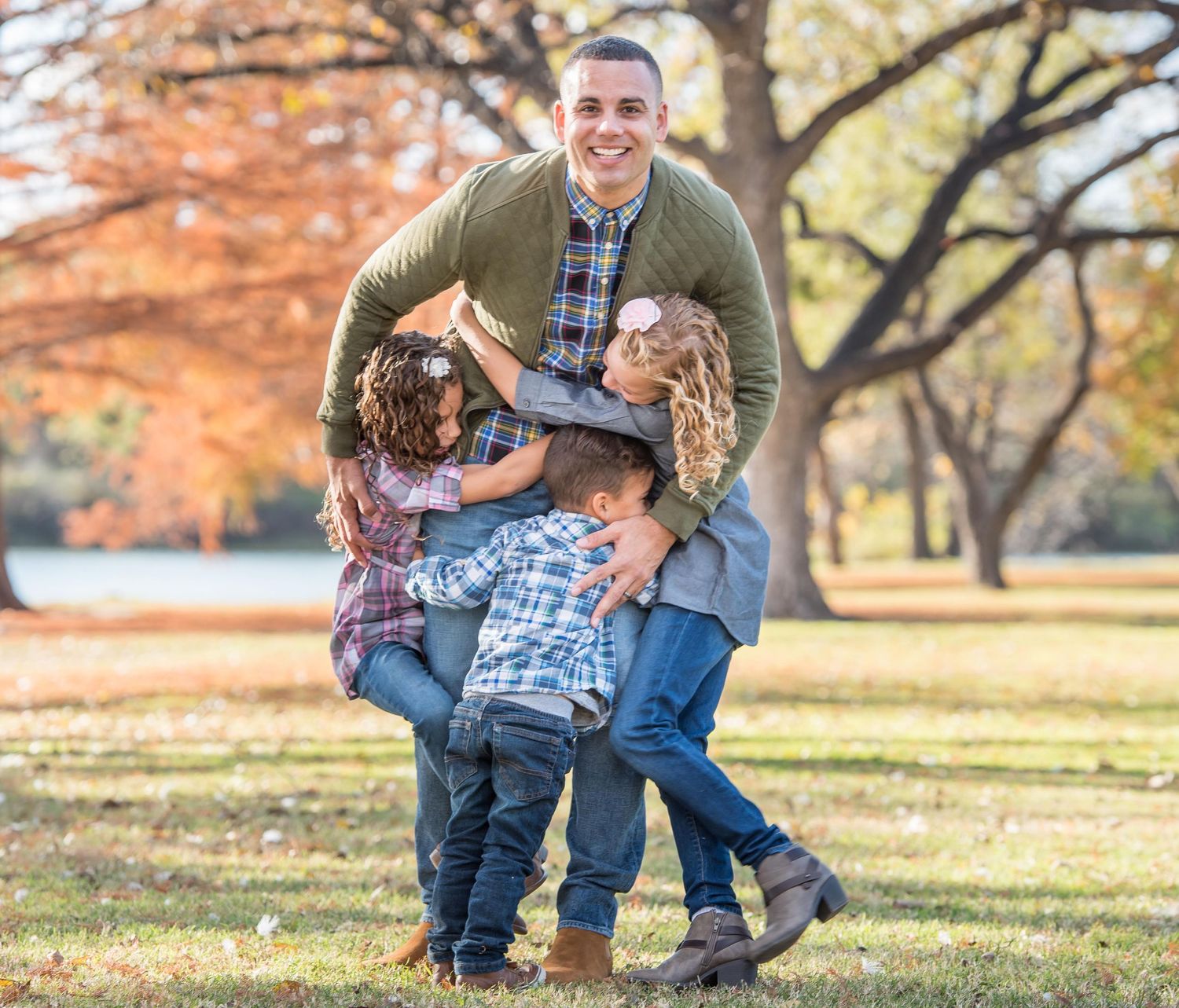 Person in olive sweater plays and laughs with children on grass in a fall park setting taken by family photographer in Omaha.