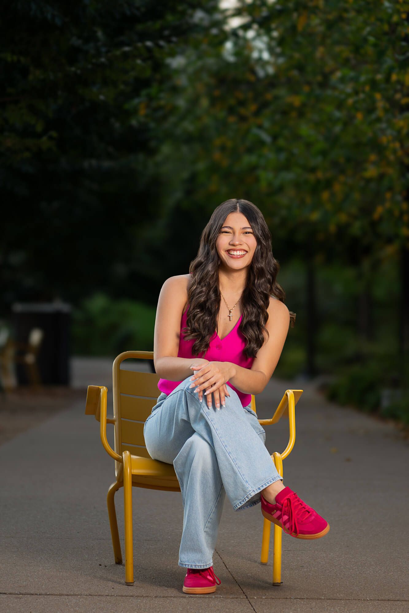 Young woman in pink top, jeans, and red sneakers smiling on a yellow bench in a park.