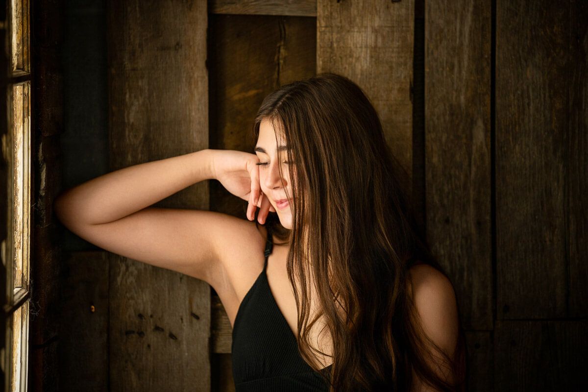 Young woman with long brown hair in black top poses thoughtfully against rustic wooden wall in warm light.