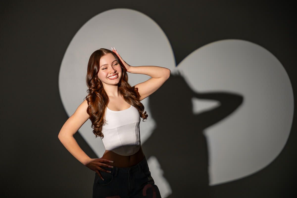 Young woman in white tank top smiling, posing with hand on hip against gray background with circular light projection.